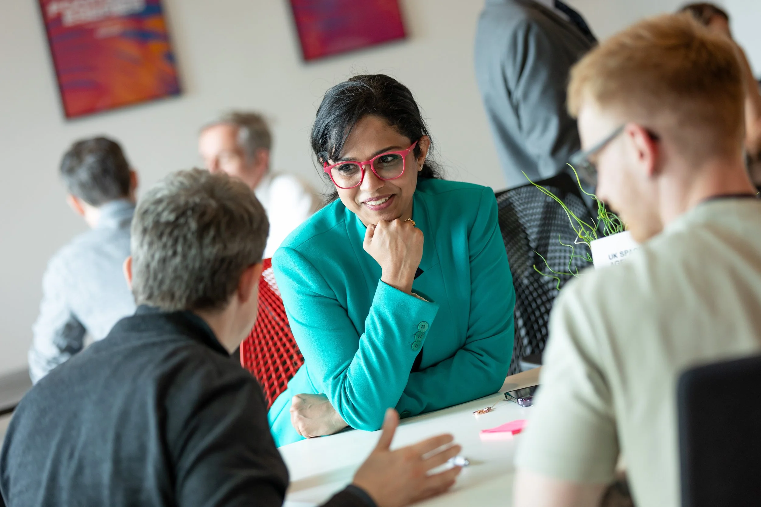A woman with dark hair, wearing glasses and a teal blazer, engaging in a discussion with two young men in a modern office setting.