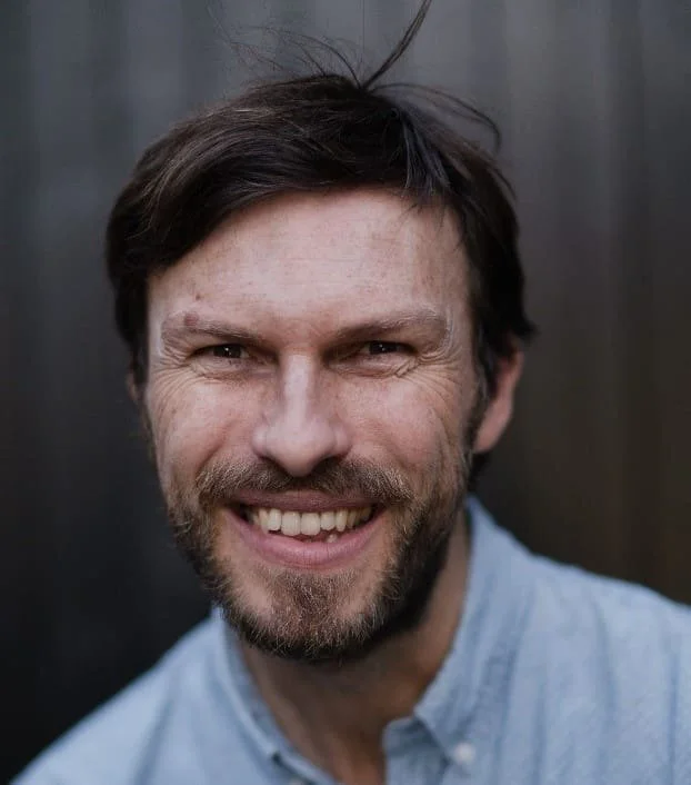 Close-up of a smiling man with dark brown hair and a beard, wearing a light blue shirt, in front of a blurred dark background.