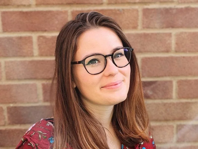 A woman with long brown hair, wearing glasses, and a maroon floral top, standing against a brick wall.