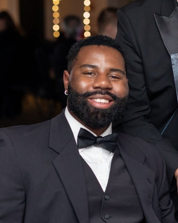 A happy man with a beard and short hair, wearing a black tuxedo with a bow tie, smiling at a formal event.