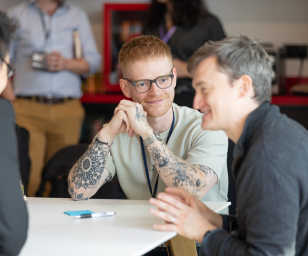 Two men sitting at a table engaged in conversation, with one man smiling and leaning on his hand, while the other talks. An office setting with other people in the background.