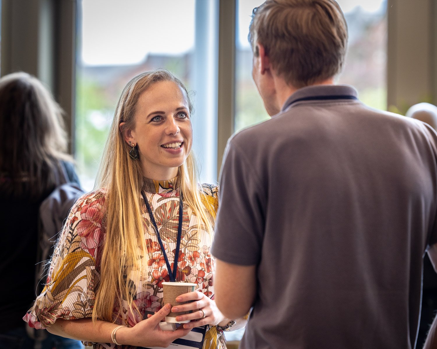 A woman with long blonde hair, wearing a floral patterned blouse, smiling and holding a coffee cup, engaged in conversation with a man whose back is facing the camera, in an indoor setting near a window.