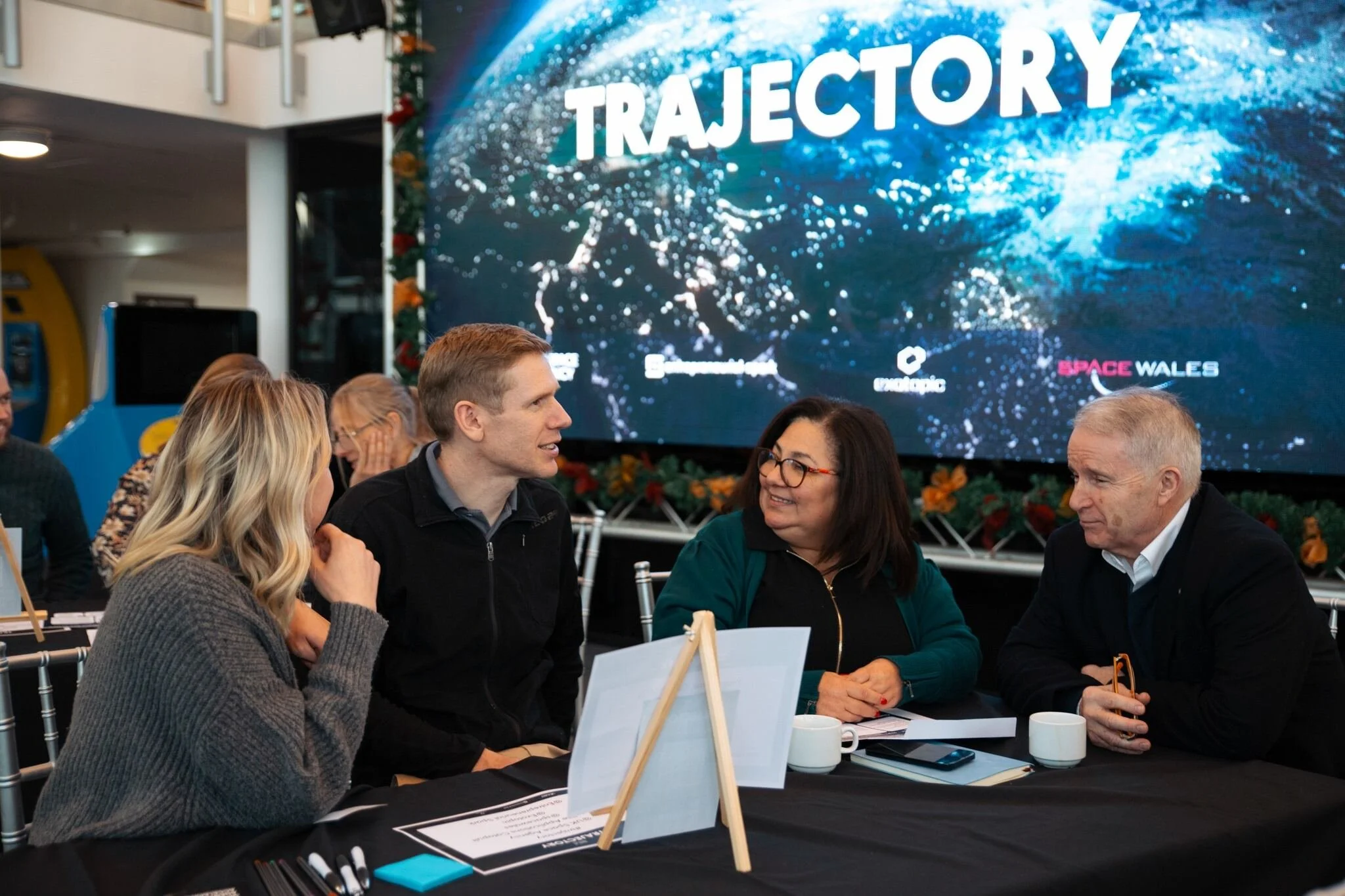 Group of people sitting at a table, engaged in conversation, at a conference or event. Behind them is a screen displaying the word 'TRAJECTORY' with a space-themed background and logos.