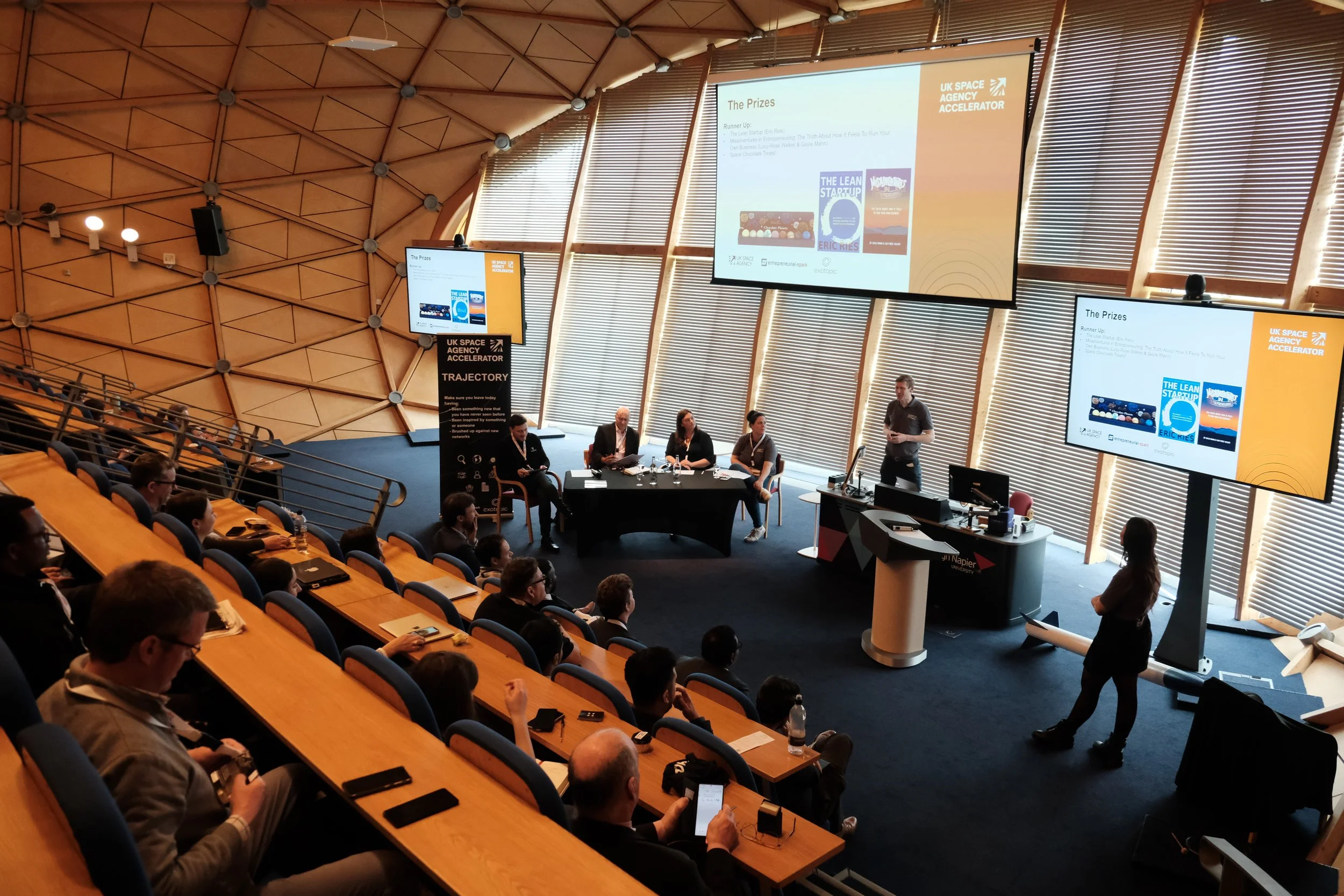 A conference room with a panel of four speakers at a table facing an audience. A presenter stands near a podium on the right. Two large screens display a presentation titled 'The Prizes' with images of books and logos, including 'UK Space Agency Accelerator'. The room features wooden walls with large vertical blinds, and attendees are seated in tiered rows, some using phones or taking notes.