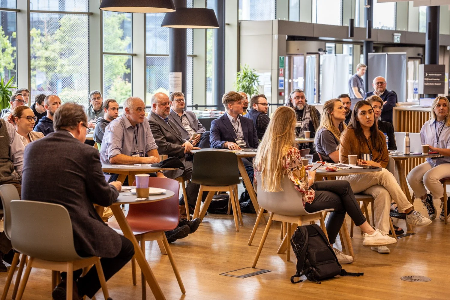 A diverse group of people attending a conference or seminar in a modern, well-lit space with large windows, sitting at tables with drinks, notebooks, and electronic devices.