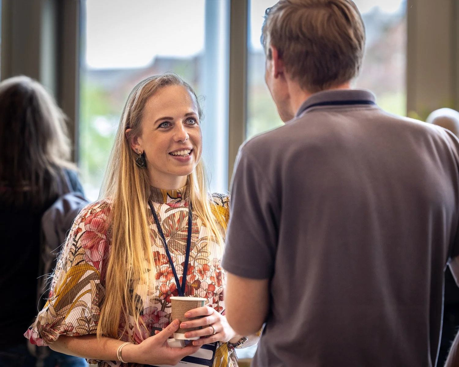 A woman with long blonde hair and floral dress smiling and holding a coffee cup, talking to a man with short brown hair wearing a gray shirt, in a bright indoor setting.