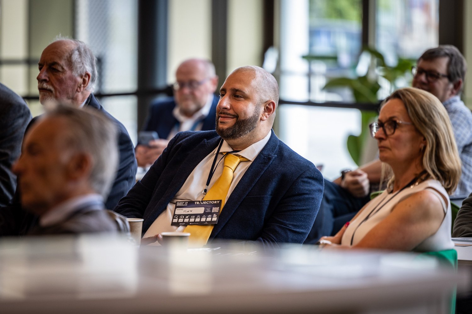 People attending a conference or seminar, sitting and listening attentively, with some taking notes or using electronic devices.