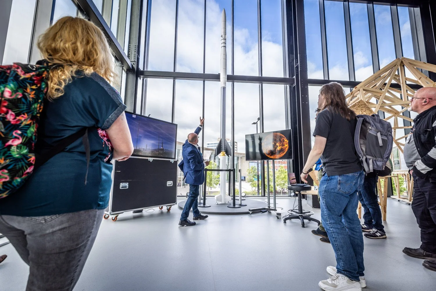Group of people observing a model rocket launch indoors, with screens displaying images of space and rockets, and a large window showing the sky outside.