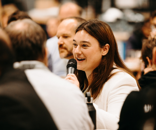 A woman smiling while speaking into a microphone at a lively indoor event.