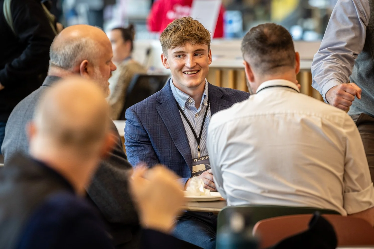 Young man in a blue blazer smiling and engaging in conversation at a group event or conference.