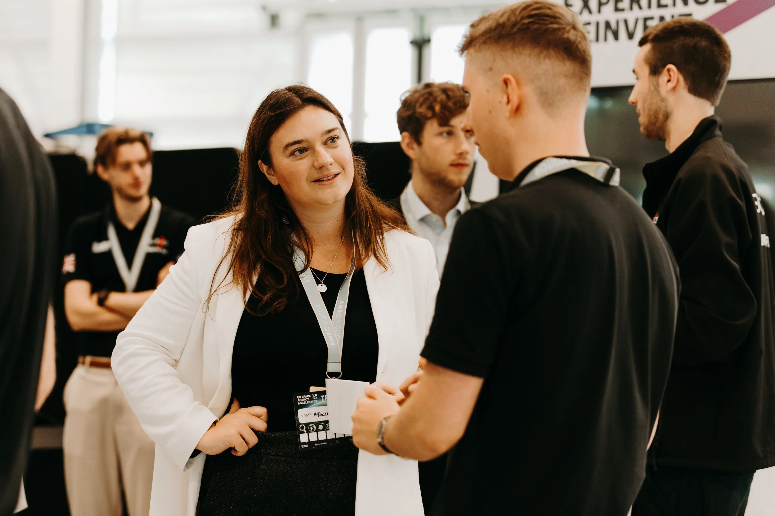 A woman in a white blazer and black top is talking with a group of young men at a professional event or conference.