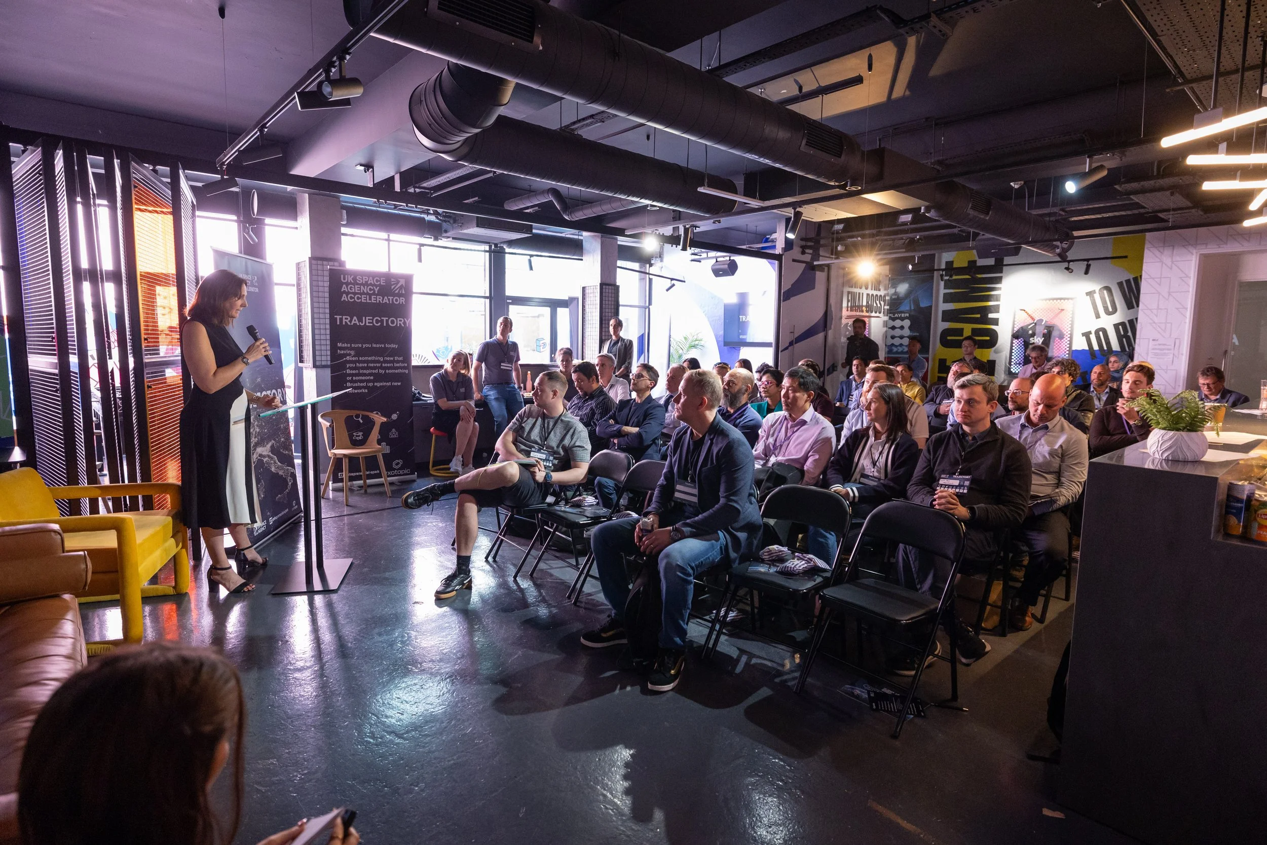 A woman in black dress speaking into a microphone at a presentation in a modern office or conference space, with an audience sitting and listening attentively.