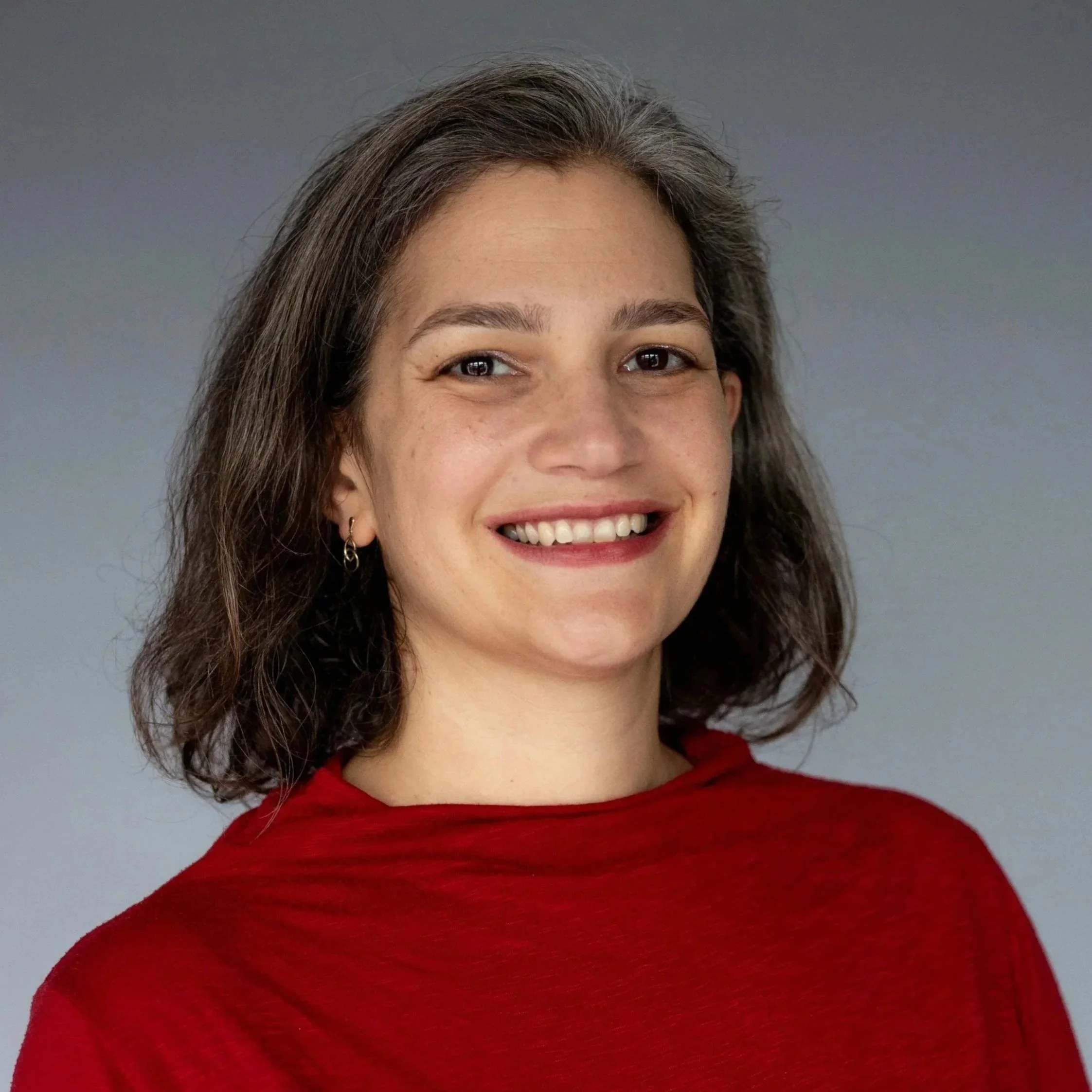 A woman with shoulder-length dark hair, wearing a red shirt, smiling in front of a plain gray background.