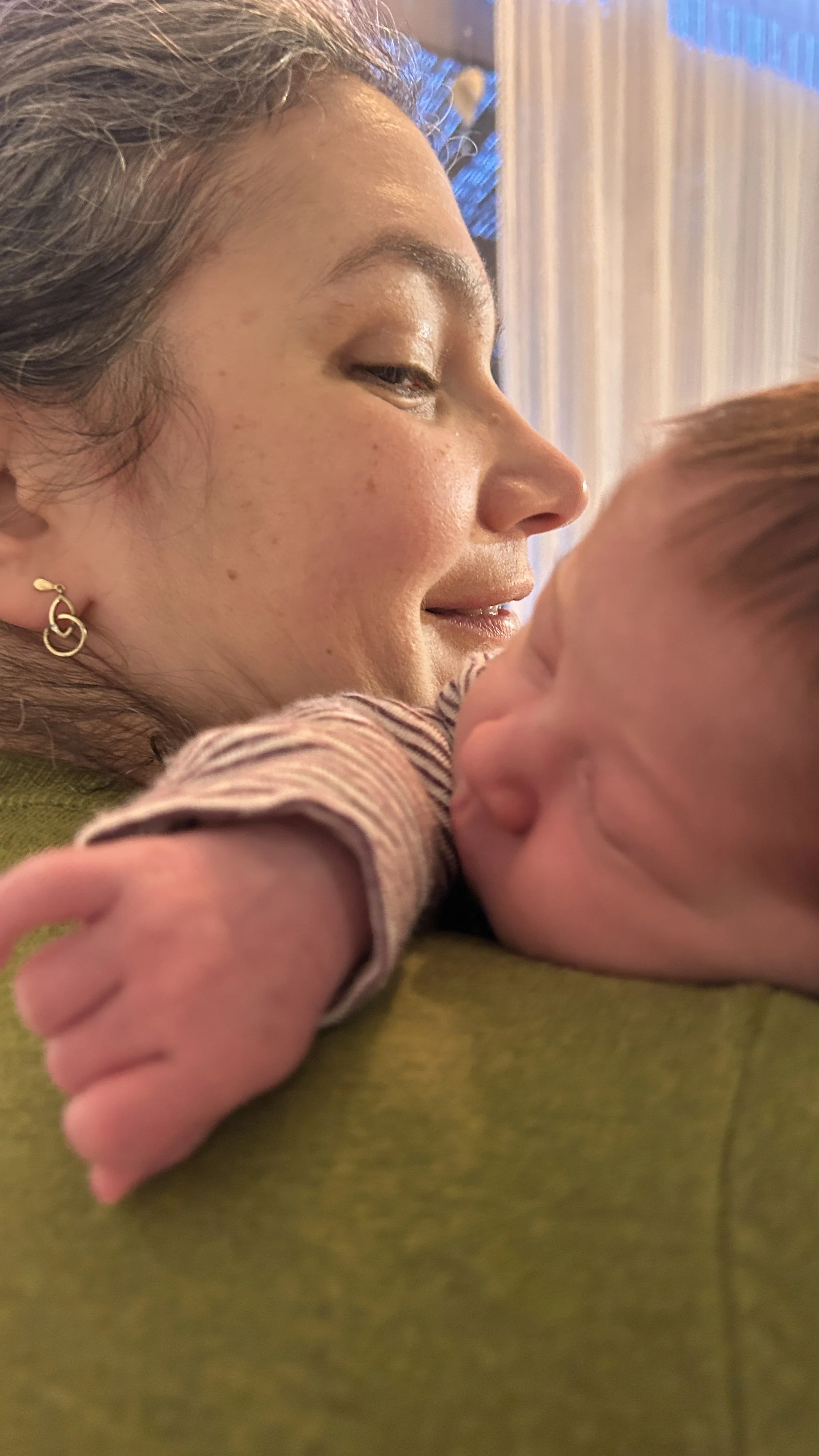 A woman with gray hair and earrings holds a newborn baby close, with both appearing to be in a tender moment.