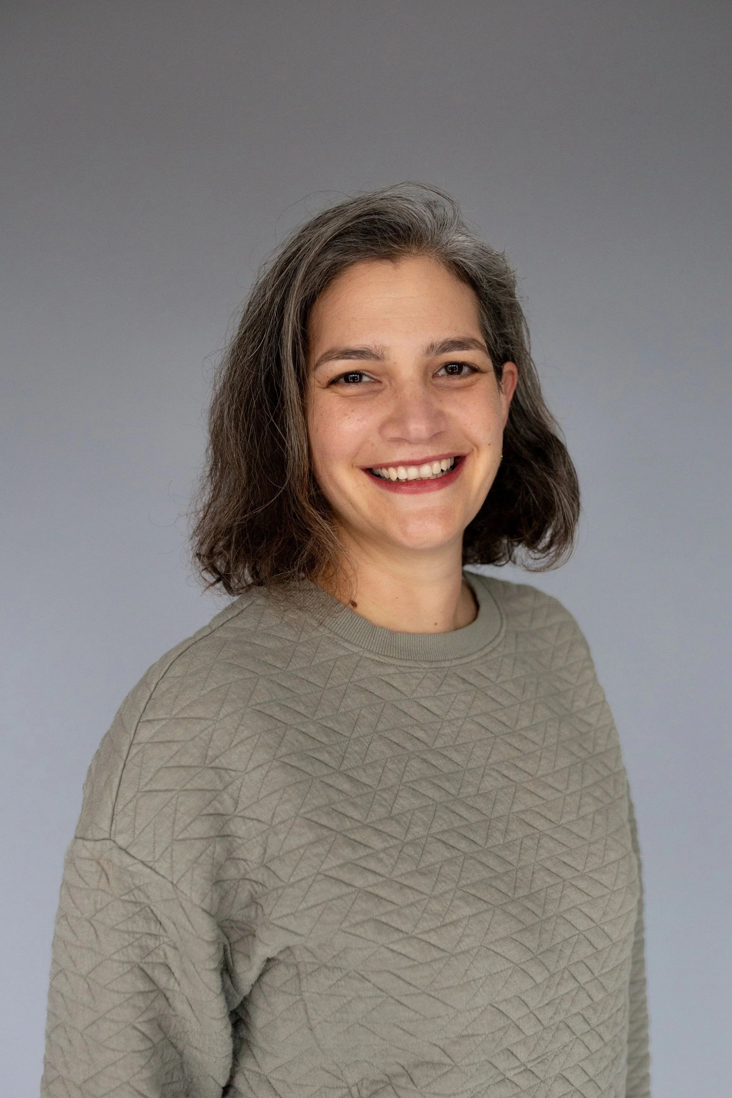 A smiling woman with shoulder-length wavy brown hair wearing a beige textured sweatshirt against a plain gray background.