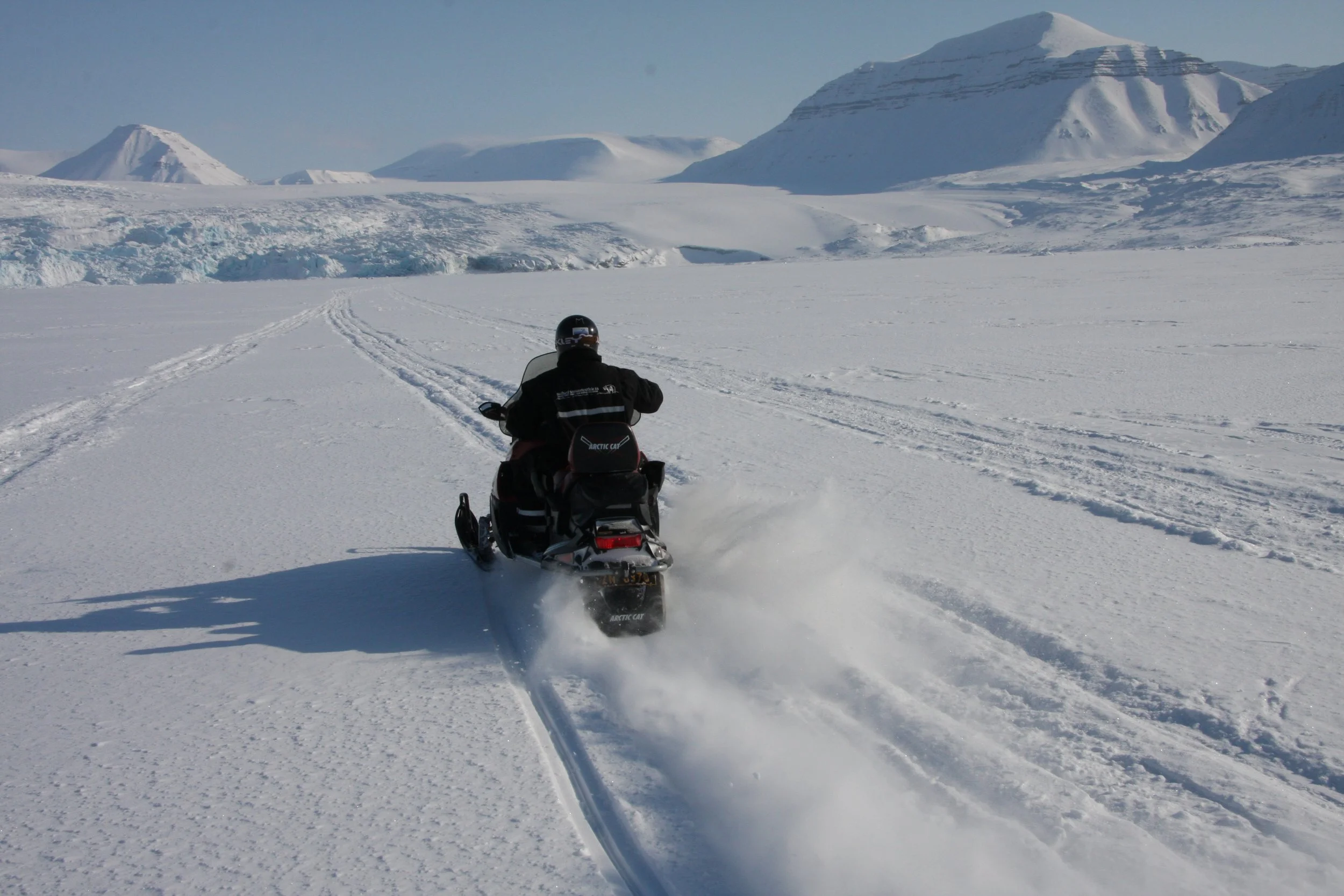 En person kjører snøscooter over snødekt landskap med snøtunge fjell i bakgrunnen på en klar dag.