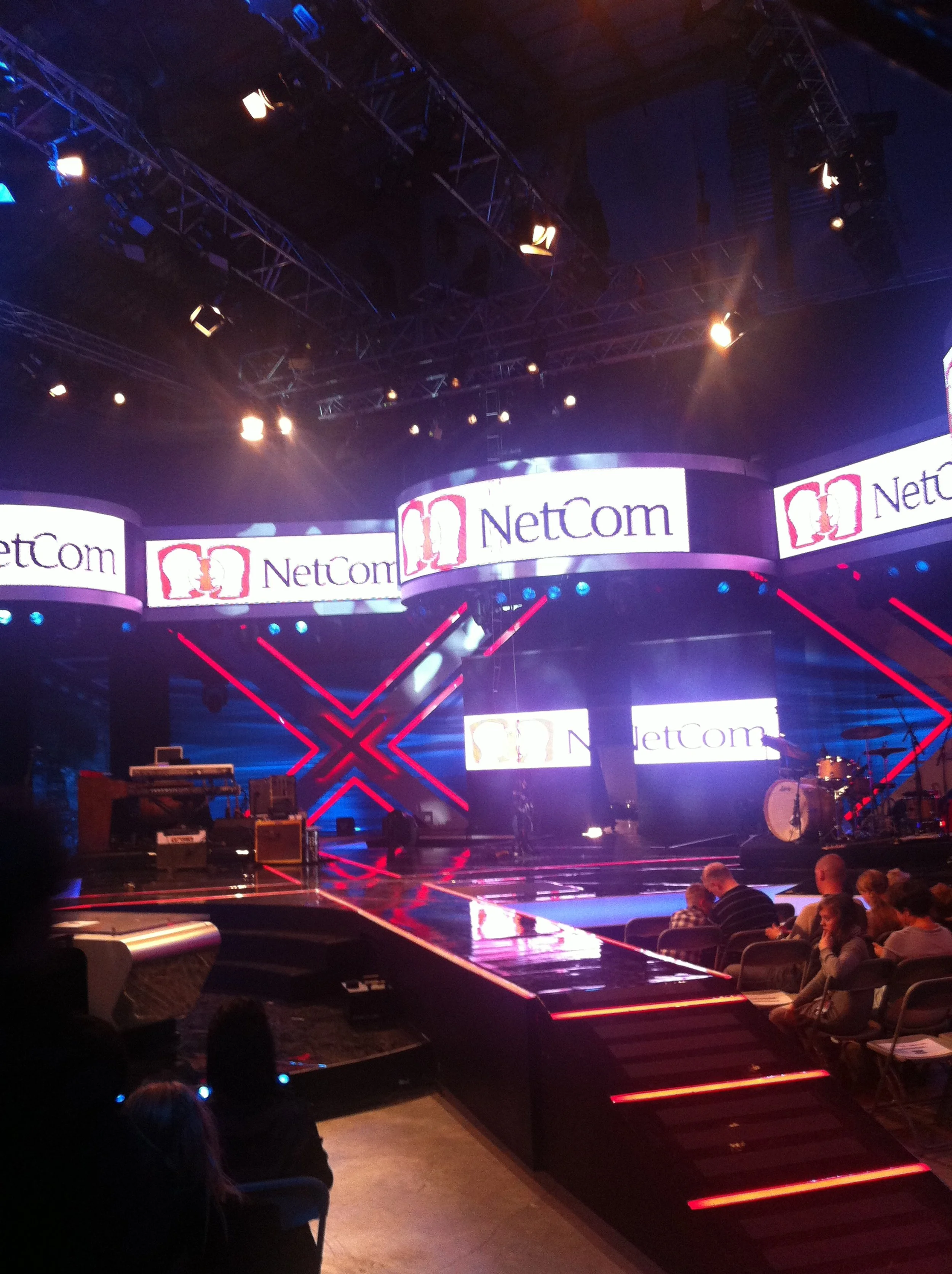 NetCom logo, og et opplyst scenegulv med noen utstyr og instrumenter, samt et publikum som sitter i forgrunn for TV 2.