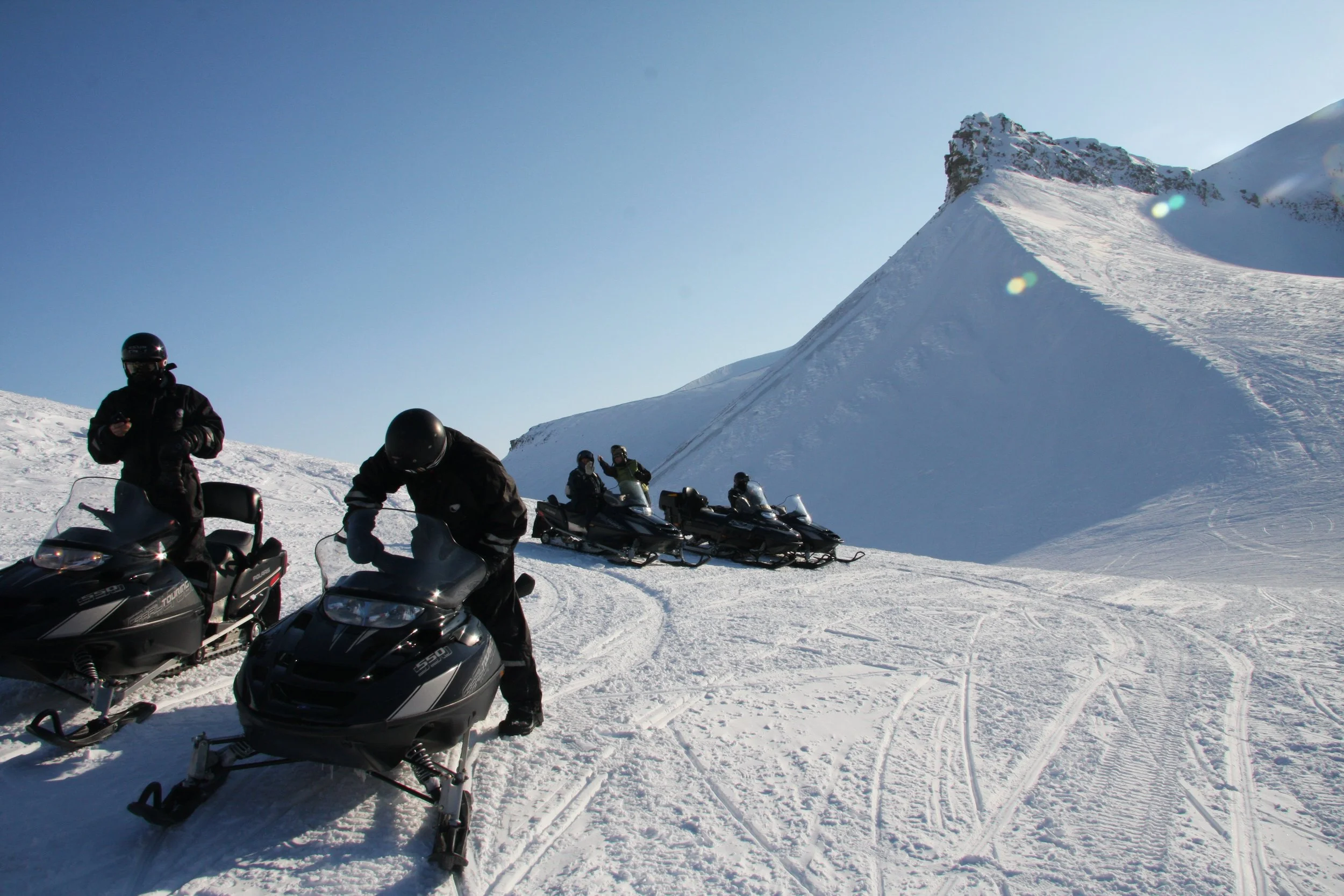 Fire personer på snøscootere i snødekt fjelllandskap under klar blå himmel.