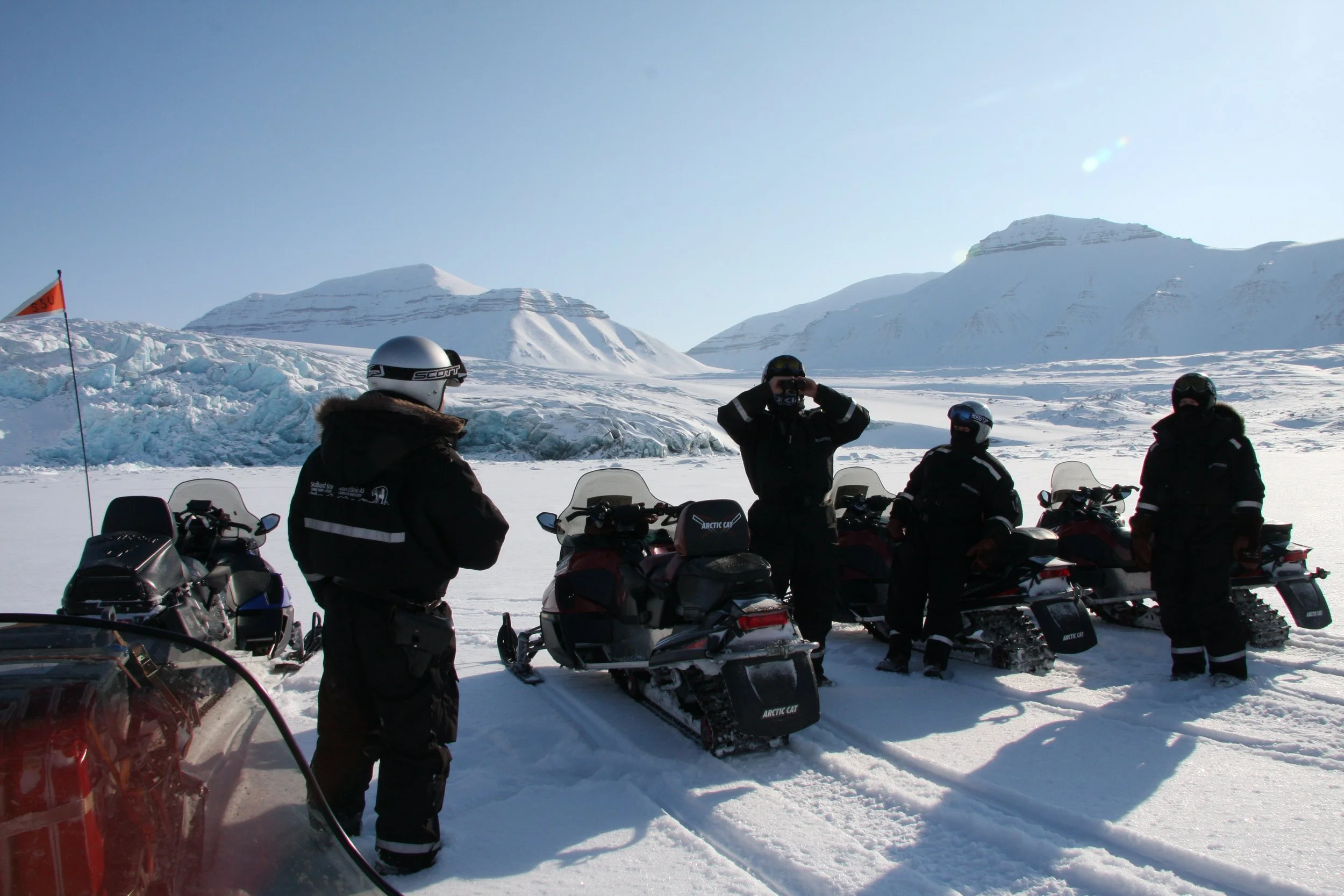 Fire personer i anorak og hjelm står ved snøscootere på snødekt landskap med snøfjell i bakgrunnen.