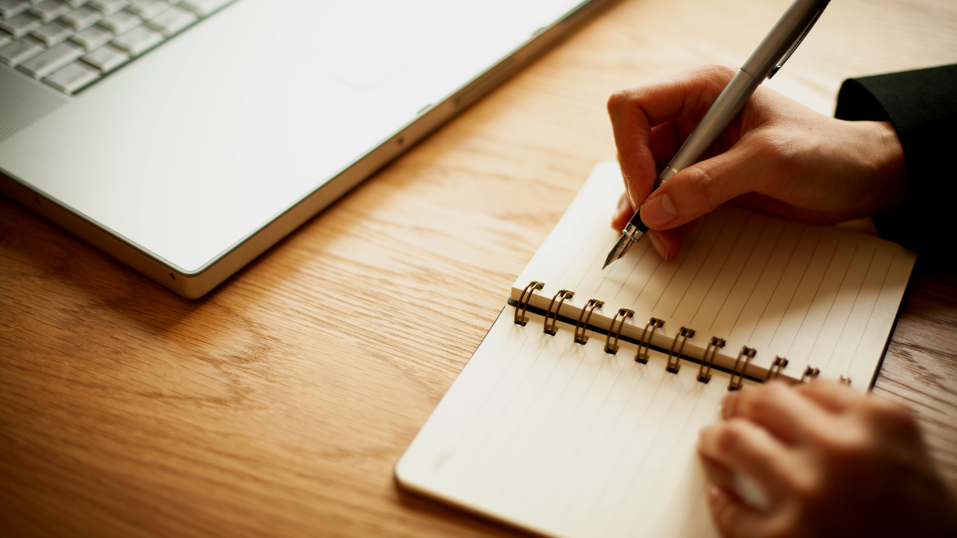 Person writing in a spiral notebook with a pen on a wooden desk, near a silver laptop with a keyboard.