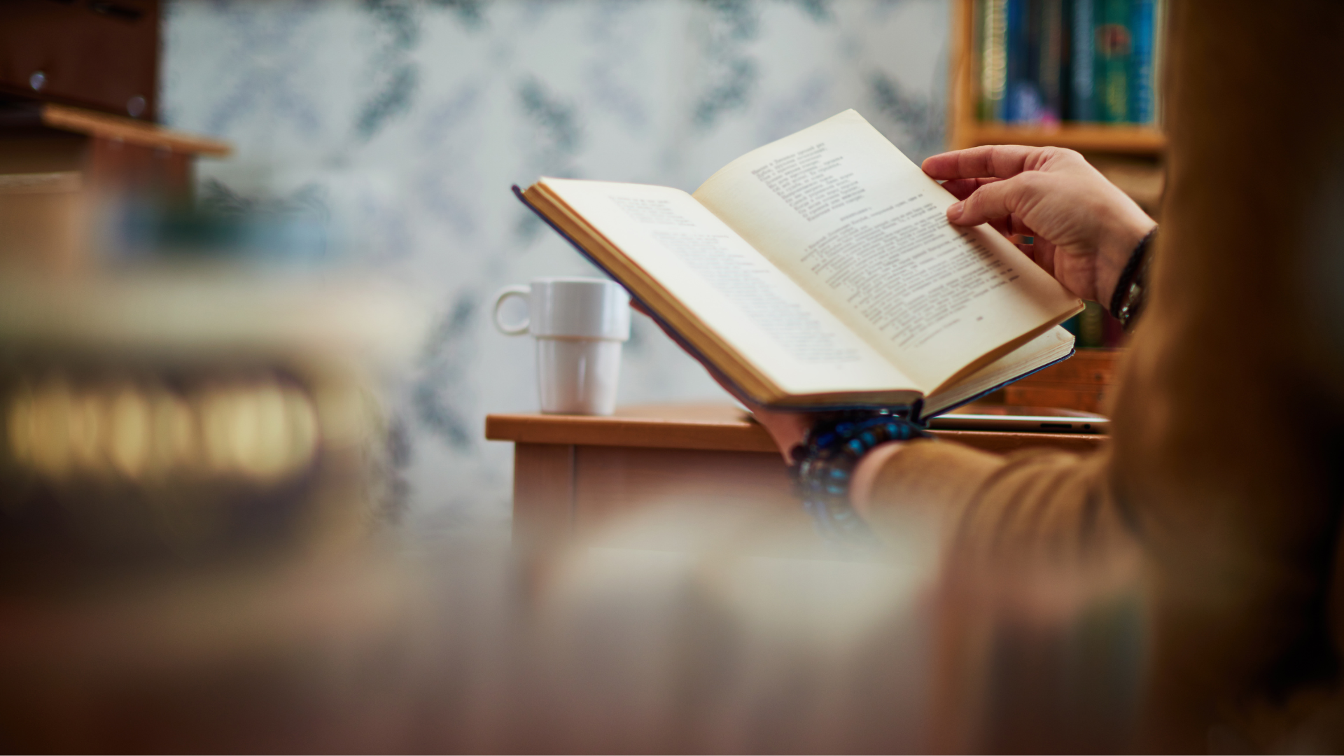 Person reading a book, mug on the desk, bookshelves in the background, and a blurred foreground.