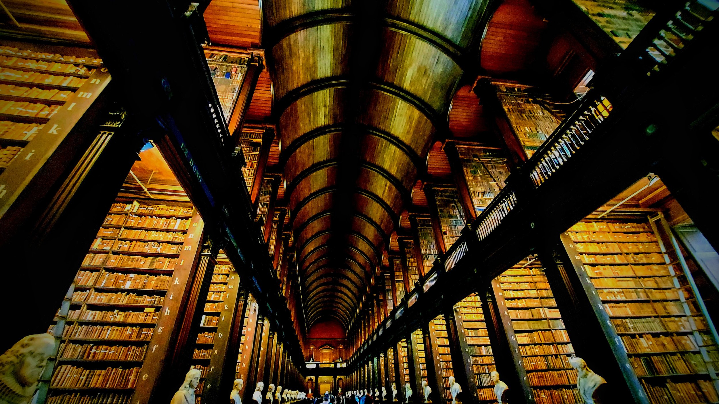 Interior view of a grand, multi-level library with tall wooden bookshelves filled with books, a vaulted ceiling, and a statue-lined balcony.
