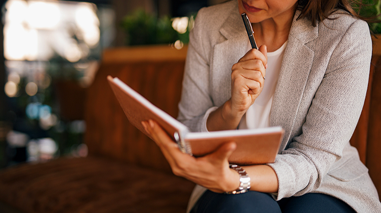 Woman in a light gray blazer writing in a notebook with a pen, sitting on a sofa in a cozy setting.
