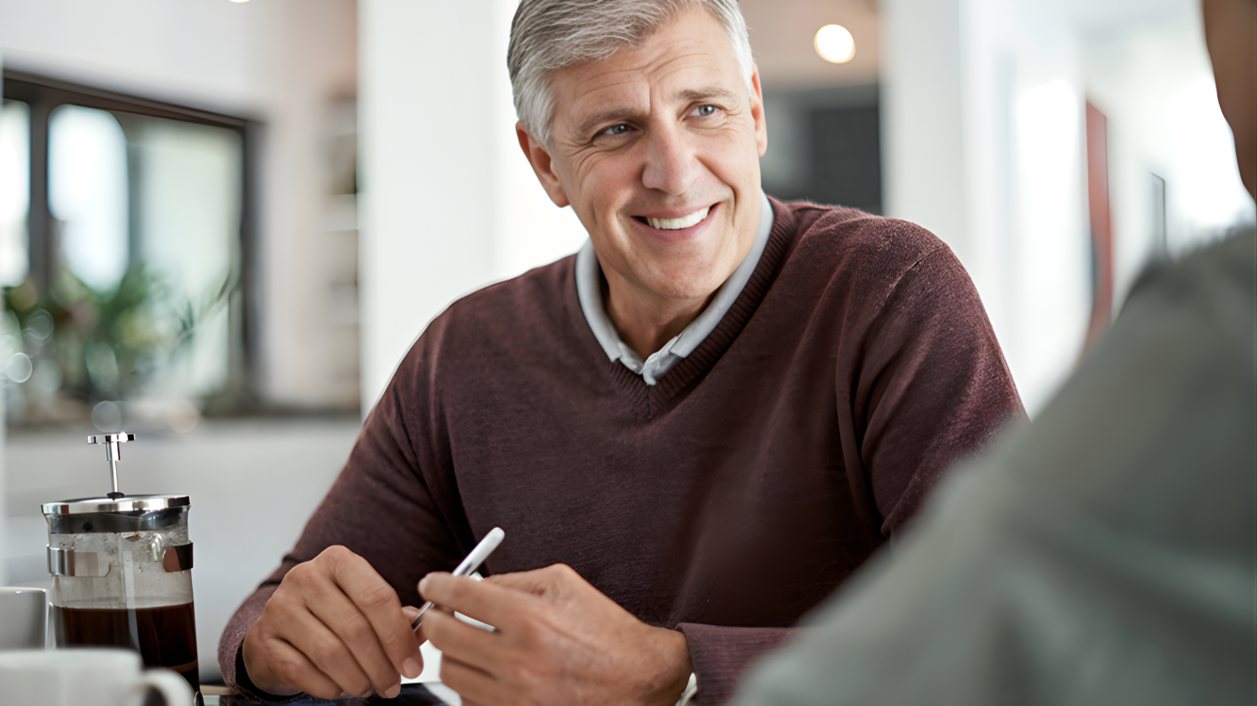 Smiling man with gray hair sitting at a table, talking to someone, with a French press coffee on the table.