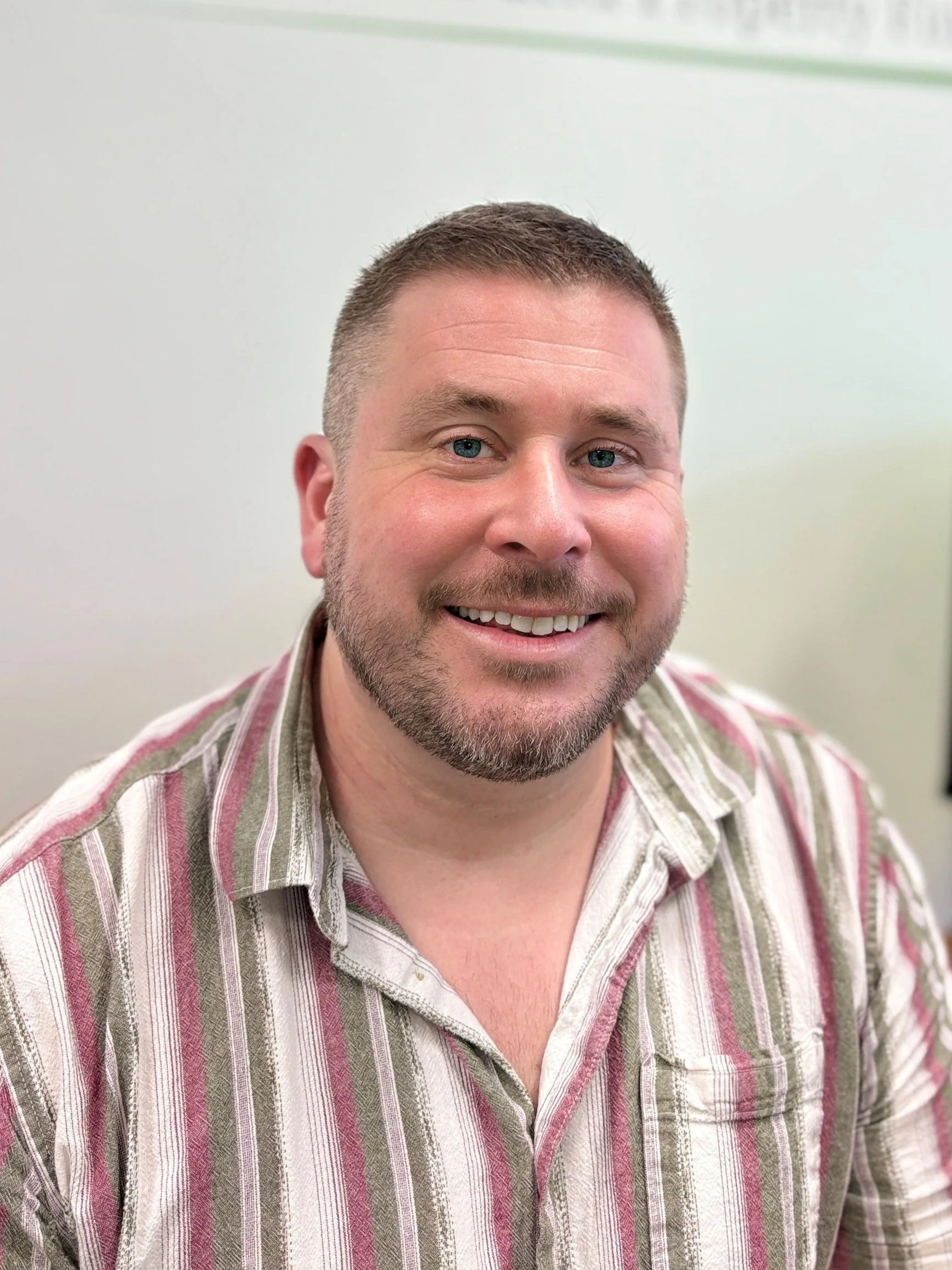 A man with short brown hair and a beard, smiling, wearing a striped beige and pink shirt, against a light background.