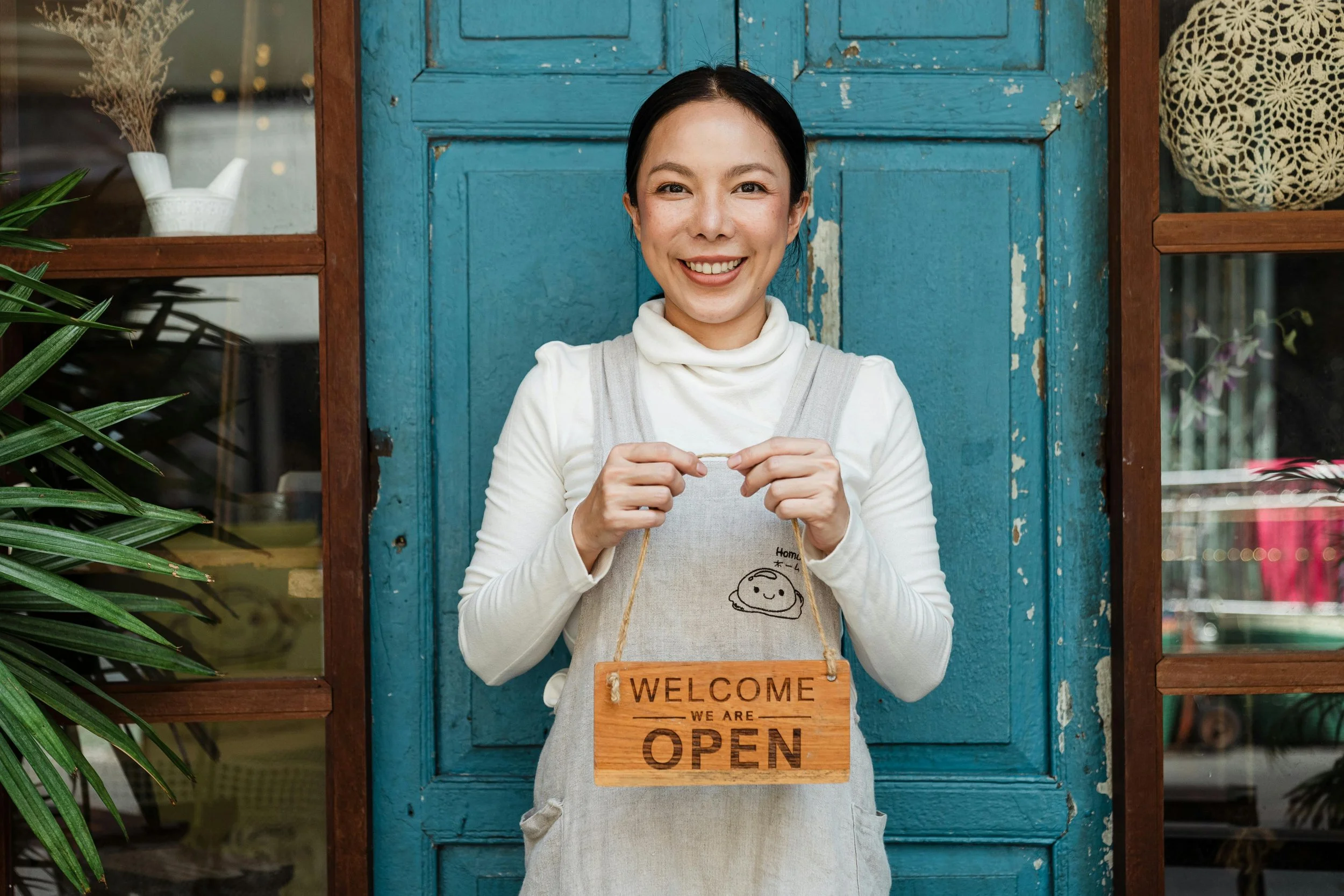 A smiling woman standing in front of a blue door holding a wooden sign that says "Welcome We Are Open"