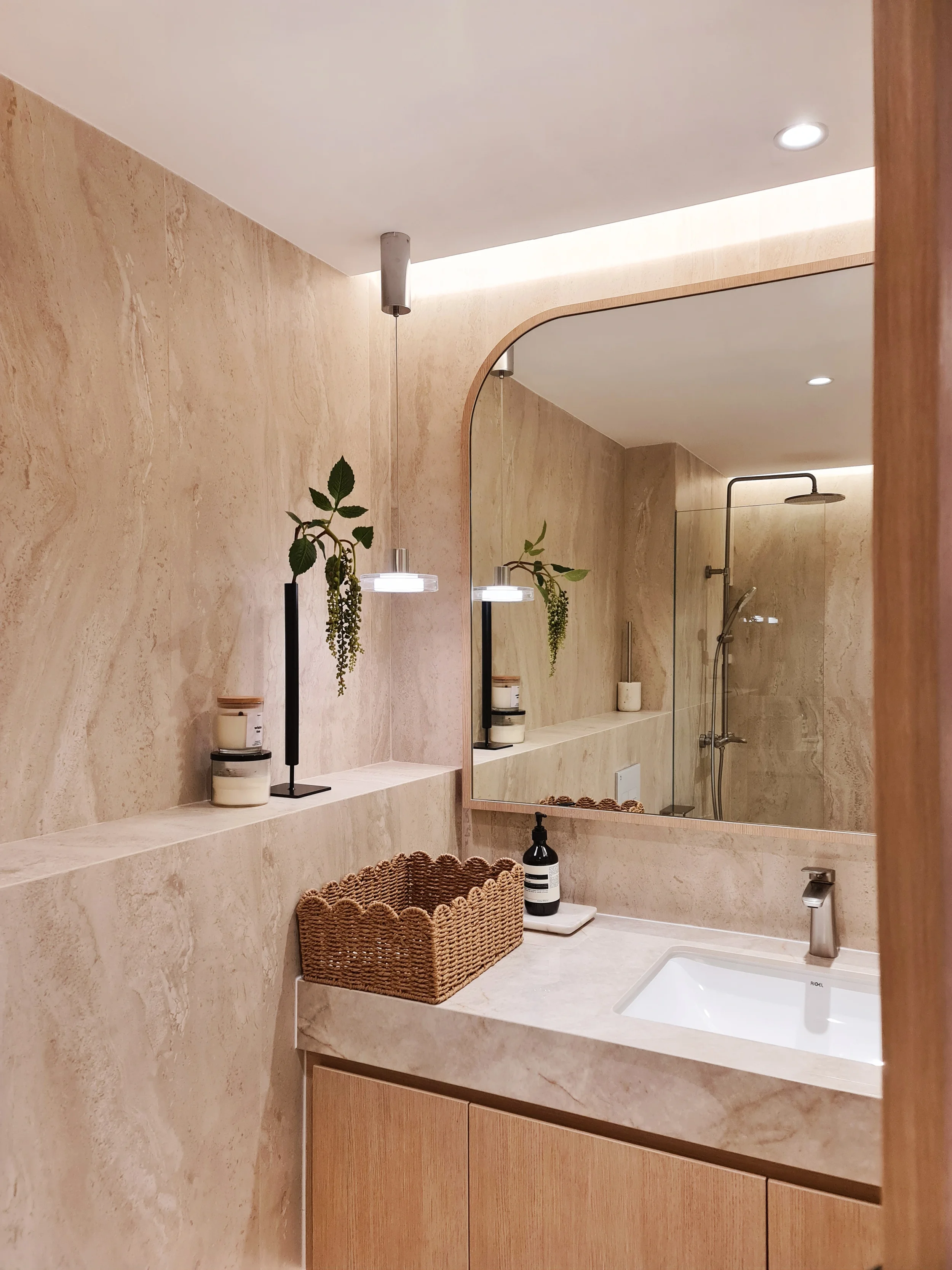 A modern bathroom with beige marble walls and a vanity area featuring a mirror, a countertop with a soap dispenser, a woven basket, and decorative plants. In the background, a shower area with a rainfall showerhead is visible.