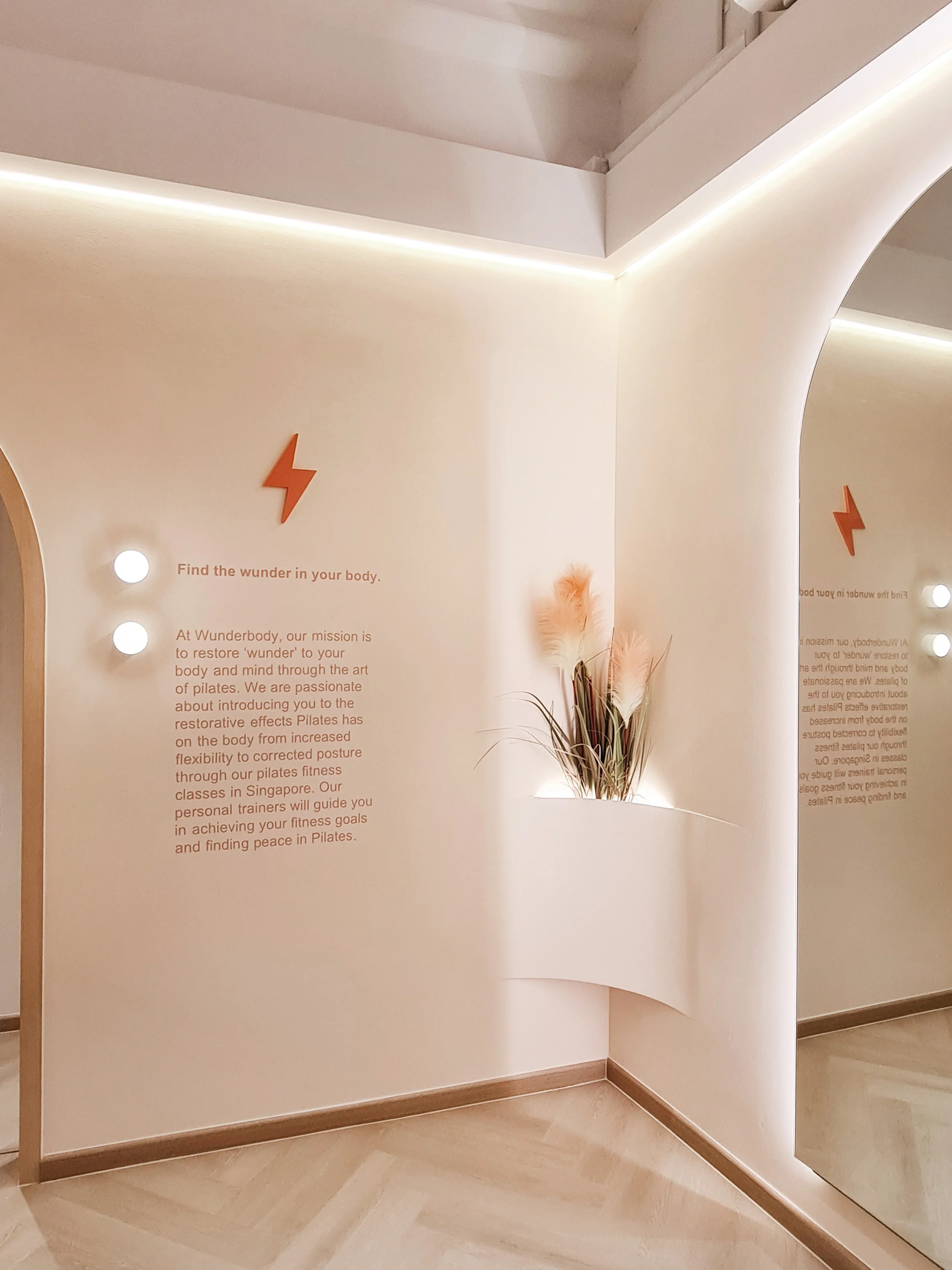 Interior corner with a small niche holding dried pampas grass, a wall with text and a lightning bolt symbol, and a mirror reflecting part of the wall. The space has a minimalist design with soft lighting.