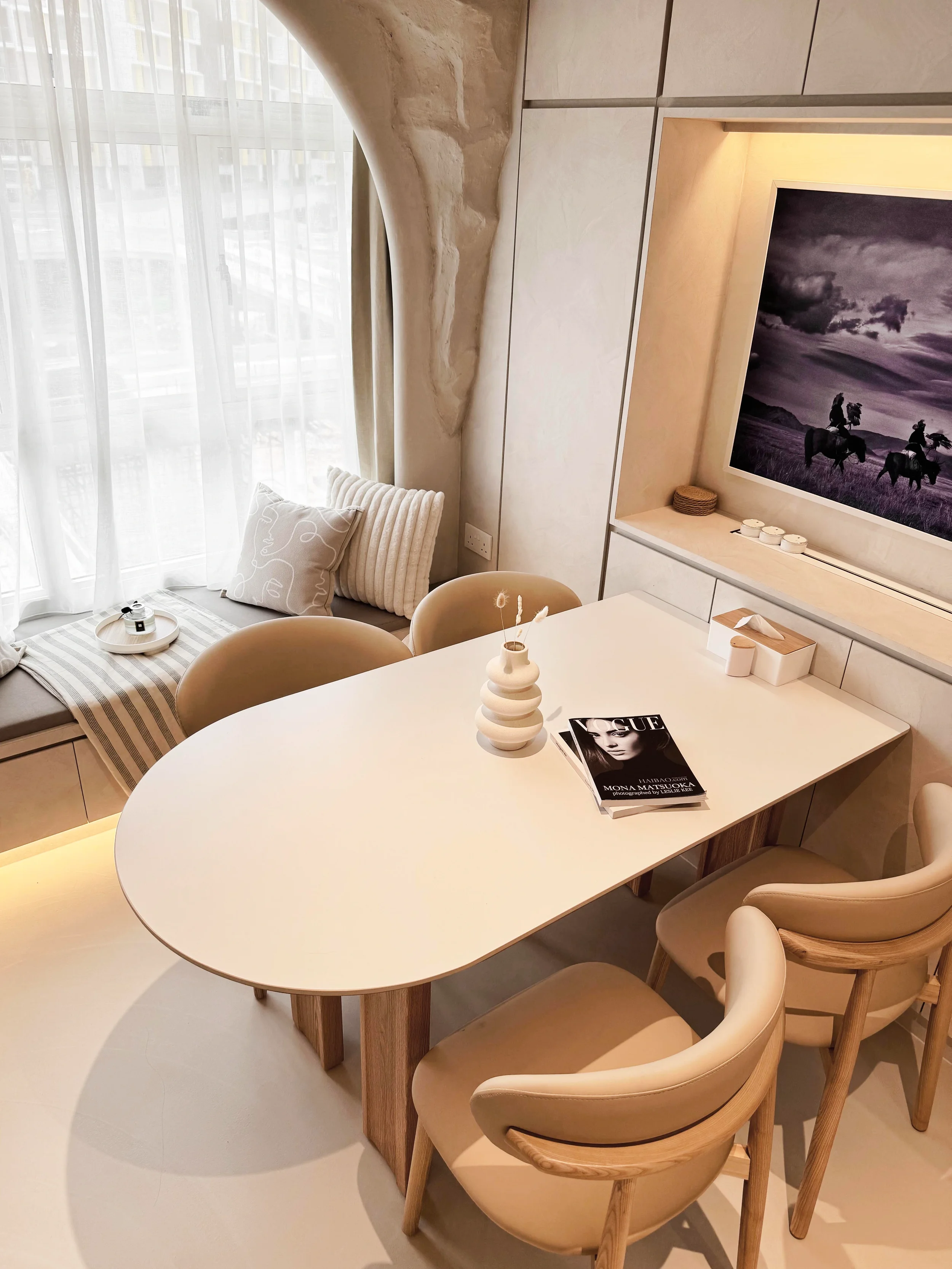 Modern dining area with an oval white table, beige chairs, and a vase with decorative sticks, set near a window with sheer curtains, and a wall-mounted TV displaying a black and white horse riding scene.