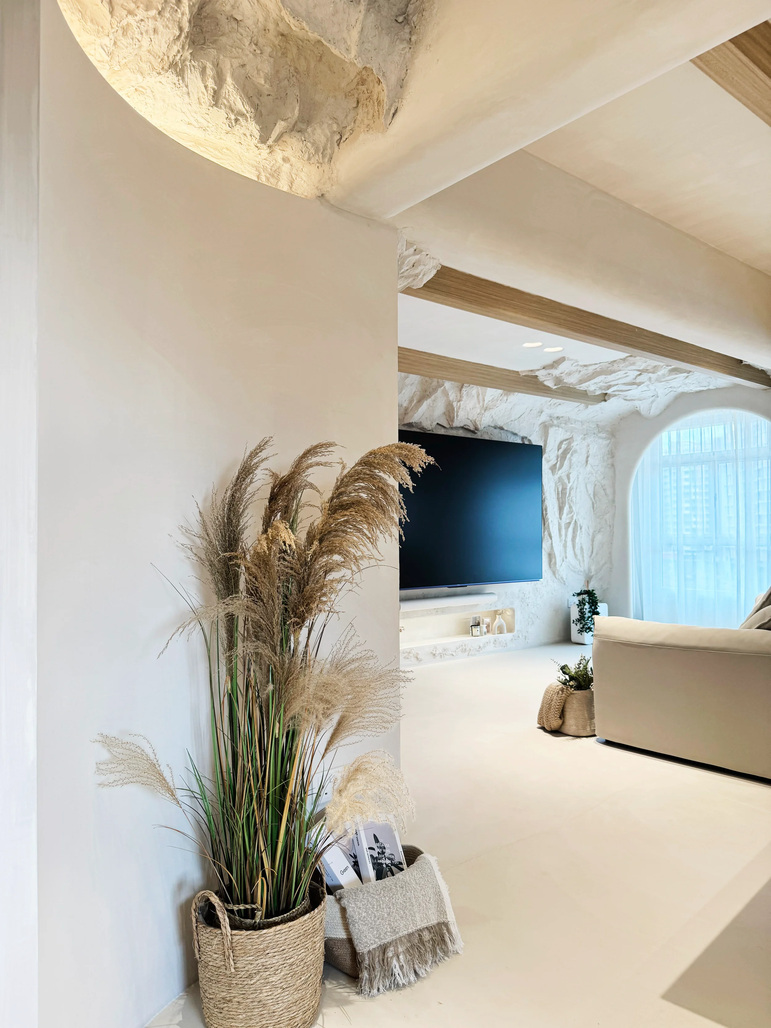 Living room with neutral tones, a large flat-screen TV on a textured white wall, a beige sofa, a basket with plants, and decorative items near a large window with sheer curtains.