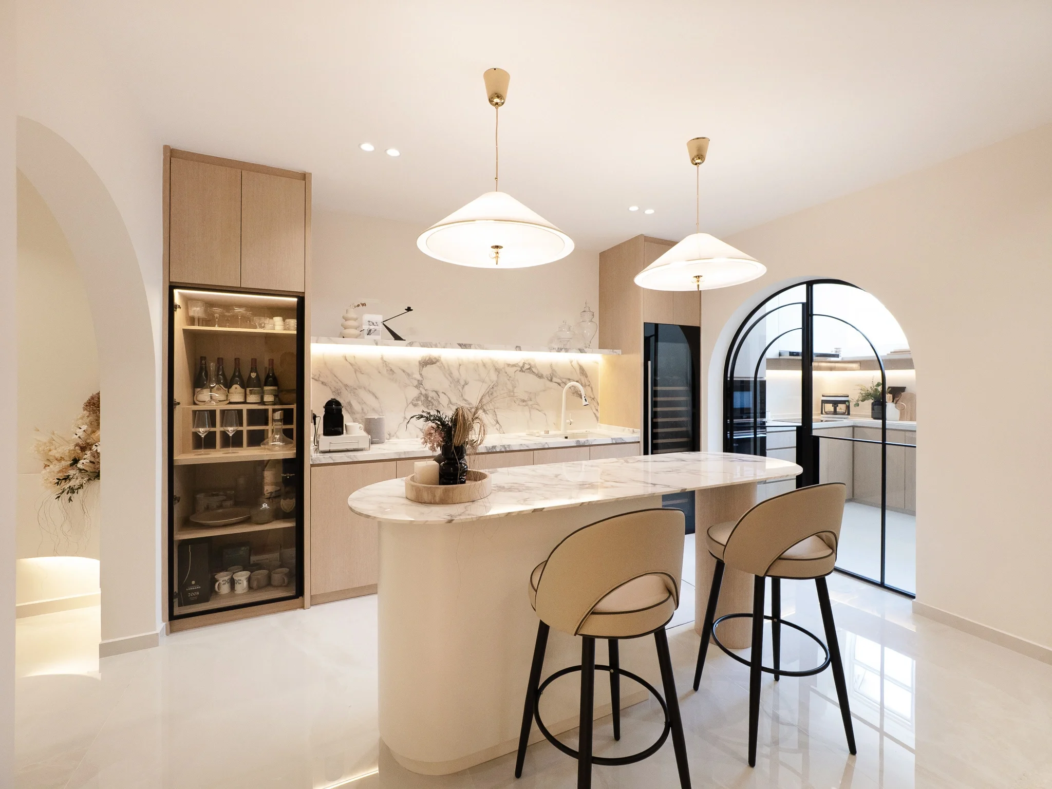 Modern kitchen with oval marble island, beige barstools, marble backsplash, pendant lights, and black-framed arched glass doors.