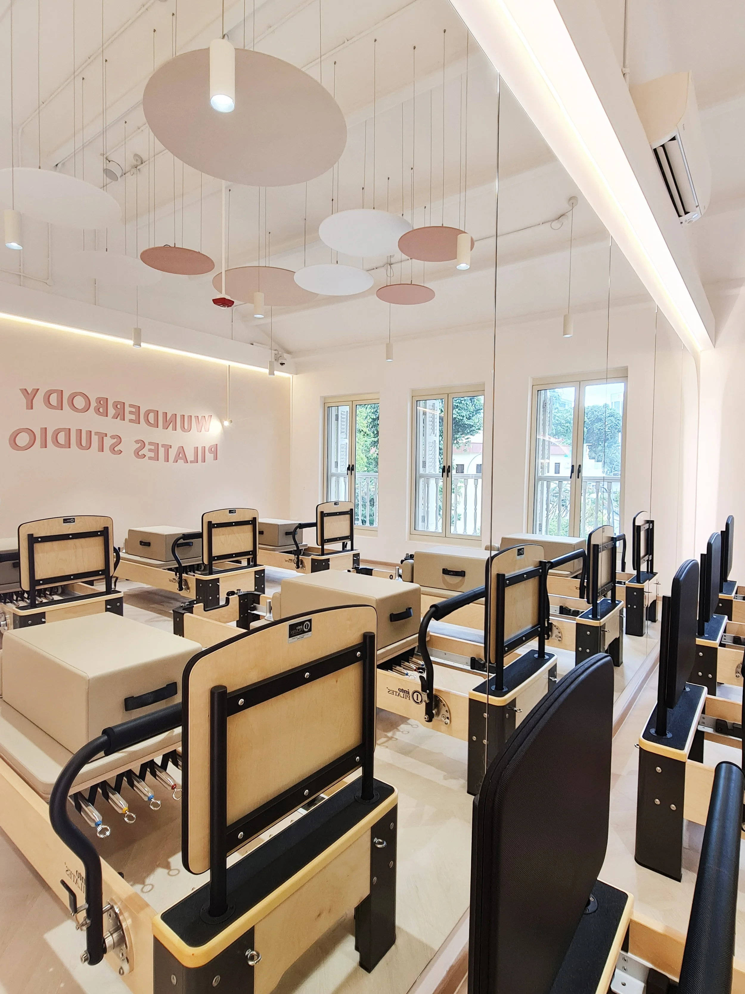 Interior of a Pilates studio with reformer machines and large windows, decorated with circular ceiling lights and a white wall displaying the words 'WUNDERBODY PILATES STUDIO'.