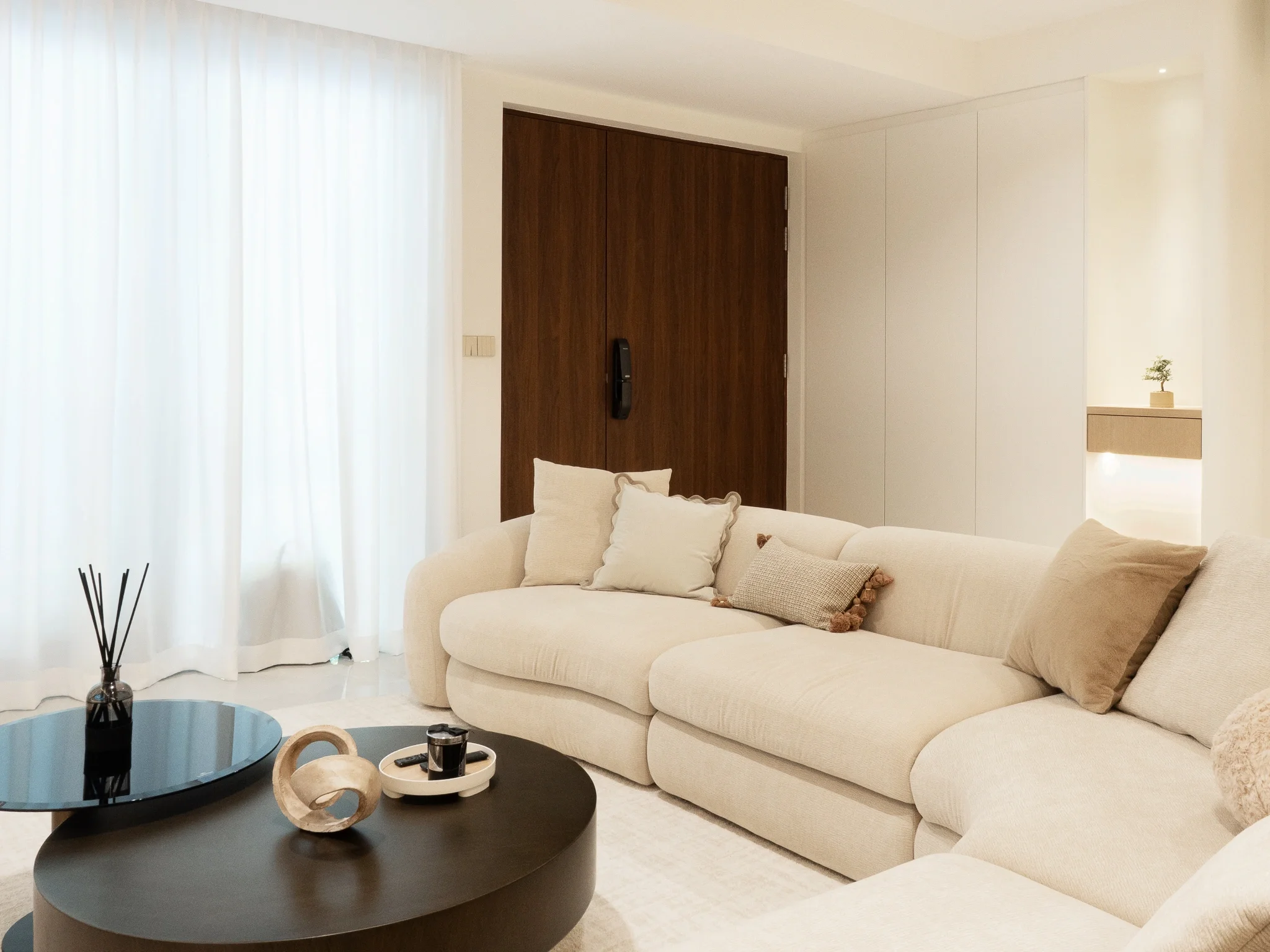Living room with a beige sectional sofa with multiple pillows, a black round coffee table with decorative items, white curtains, and a wooden door with a black handle.