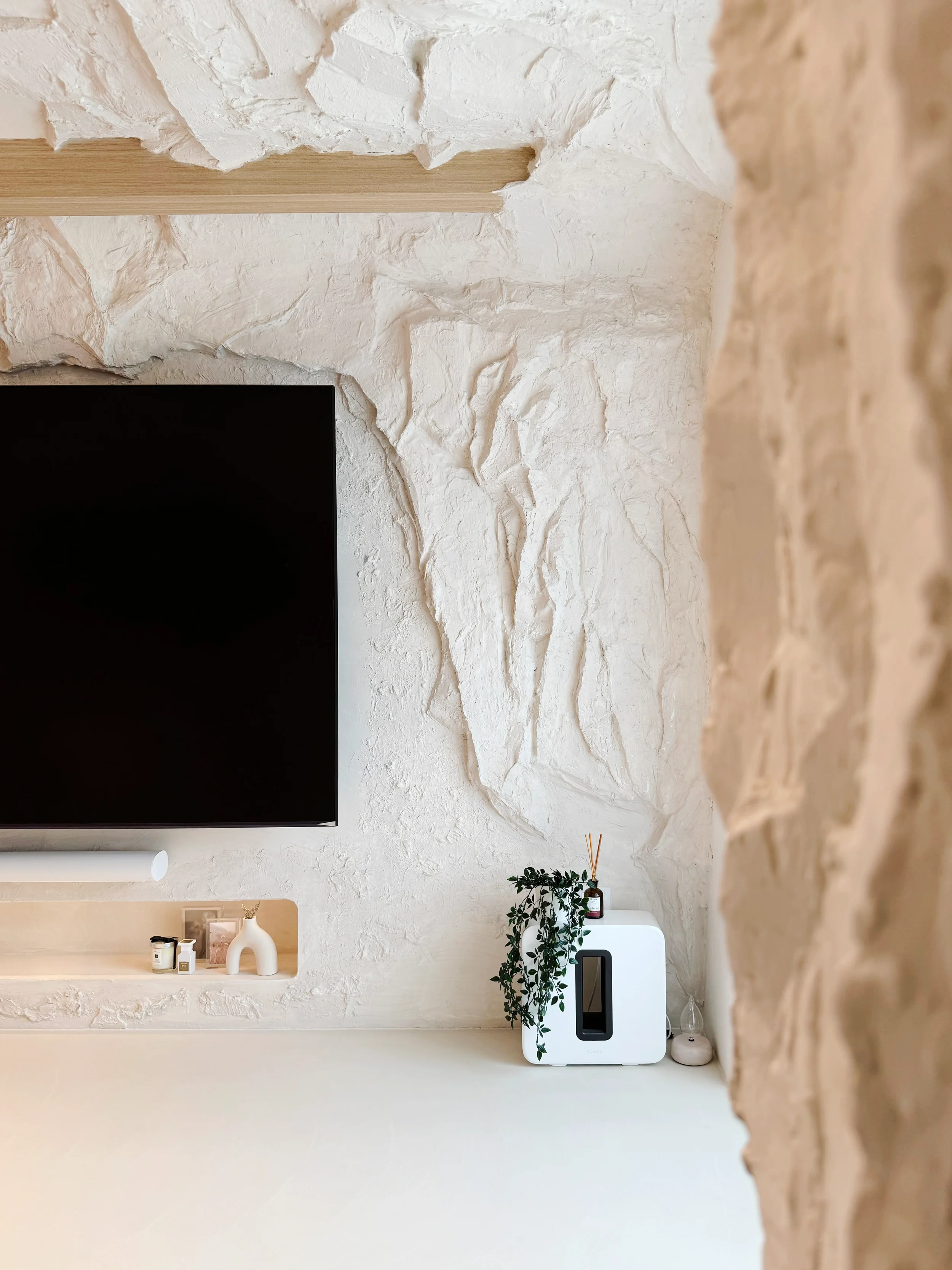 Minimalist living room corner with white textured stone wall, a wall-mounted TV, a small shelf with decorative items, and a white portable heater or speaker with a plant on top.