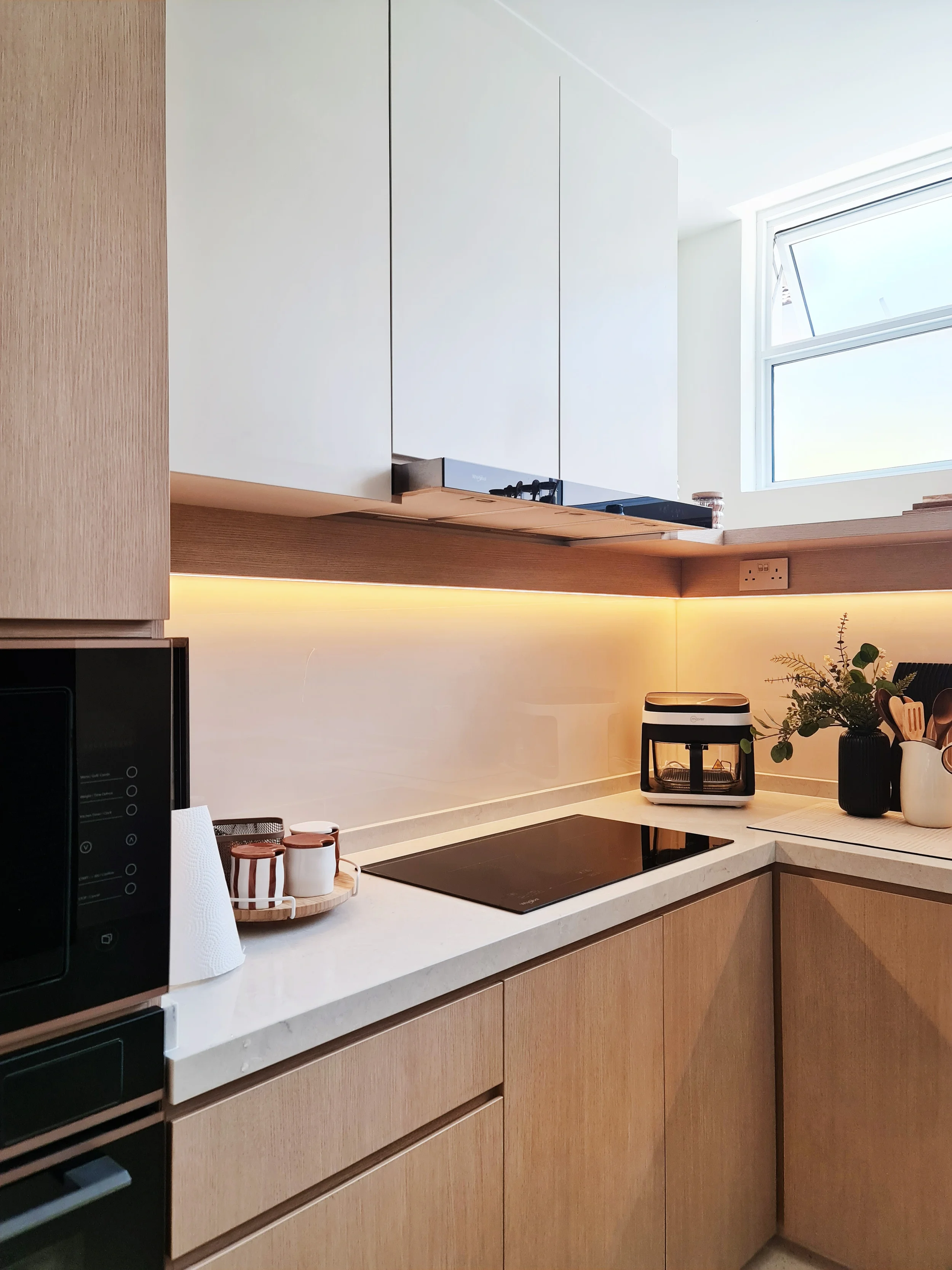Modern kitchen with white upper cabinets, wooden lower cabinets, and a marble countertop. Items include a microwave, paper towel roll, small jars, a black and white appliance, a potted plant, and utensils, with a window above offering natural light.