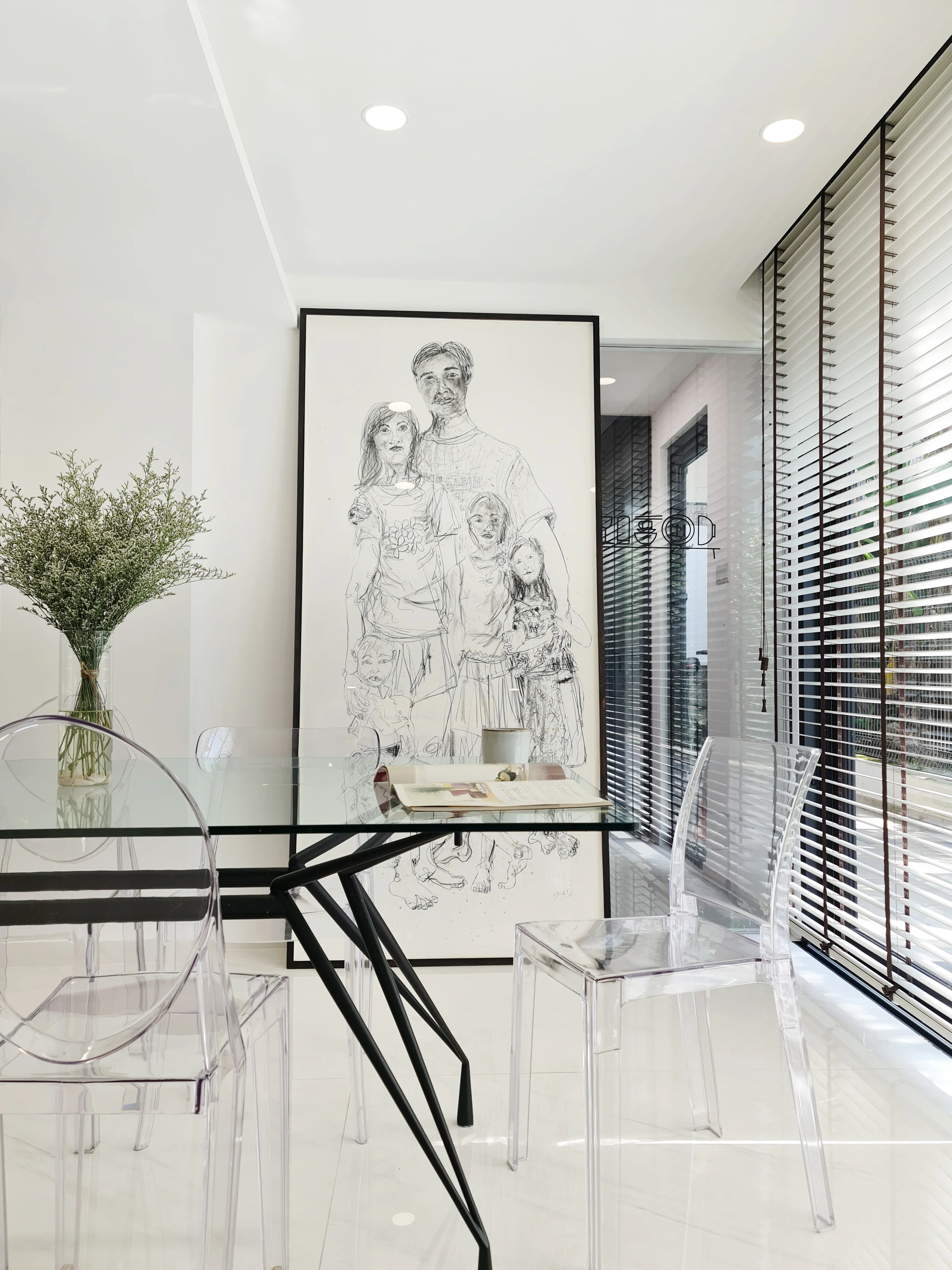 Modern dining area with a glass table and transparent chairs, a large black and white family portrait on the wall, wooden blinds on the window, and a vase with greenery on the table.