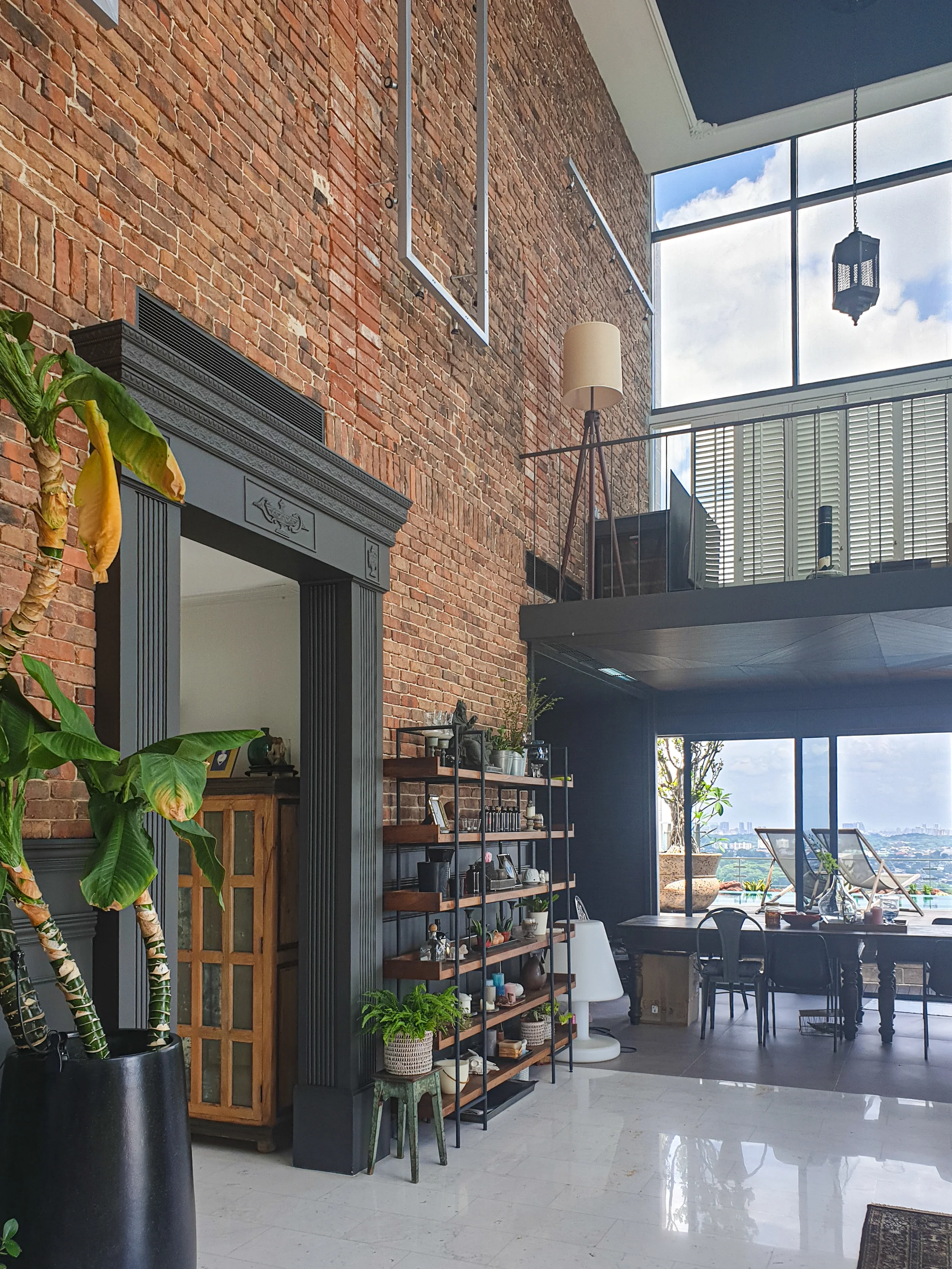 Industrial-style living room with exposed brick walls, large windows, plants, a bookshelf, and a loft area.