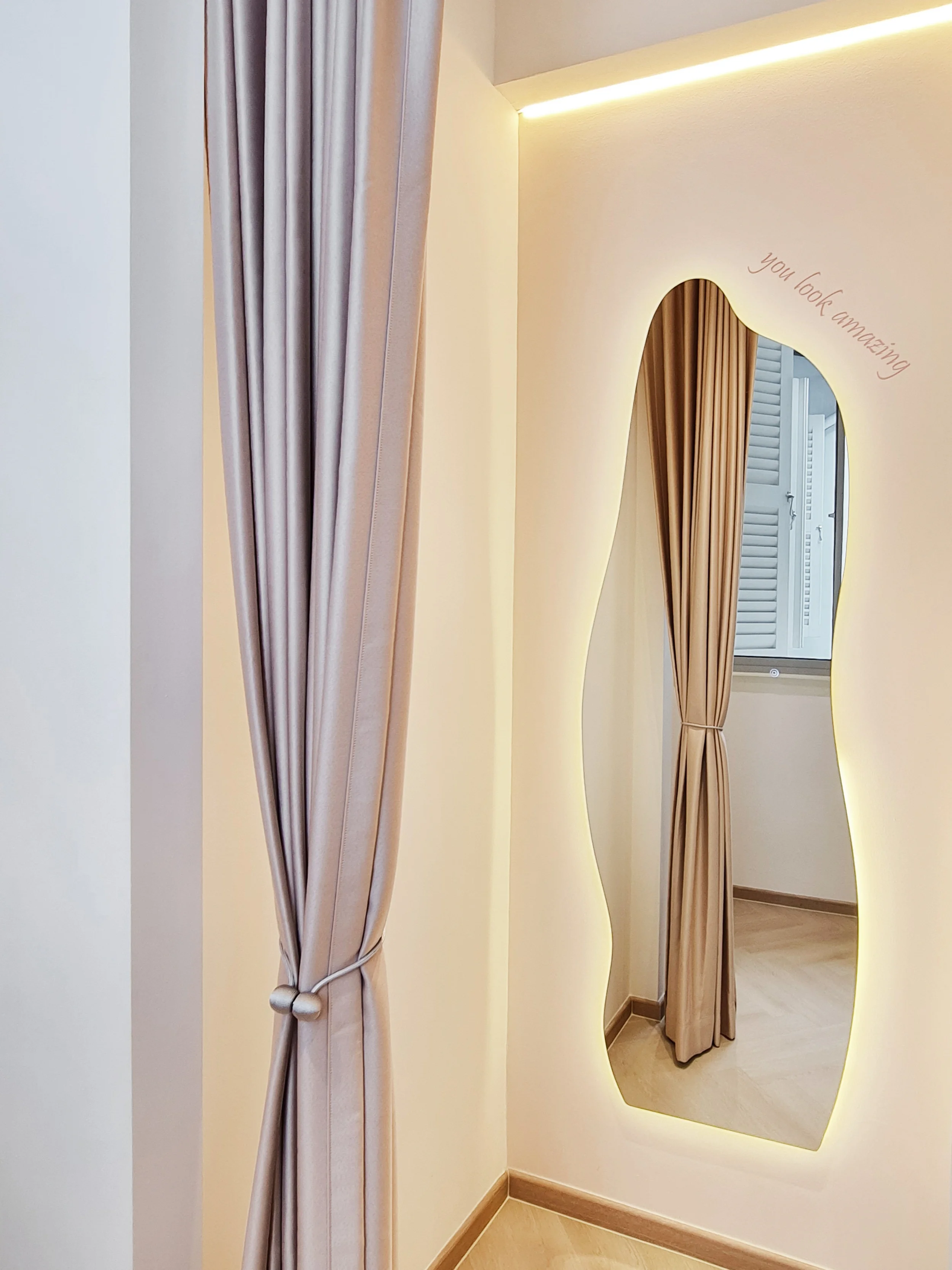 A beige curtain tied back reveals a mirror with a wavy shape on a cream-colored wall; above the mirror, a soft light and a handwritten message say, 'You look amazing'.