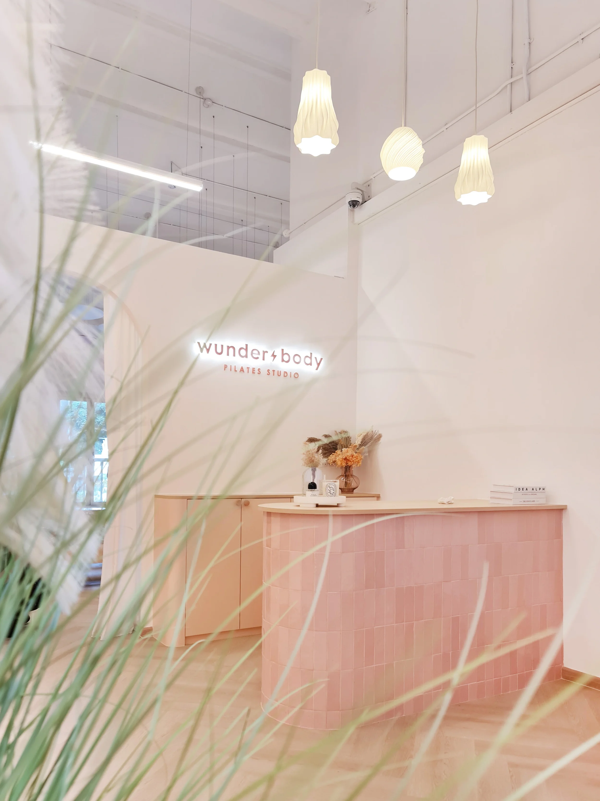 Interior of a Pilates studio with a pink curved reception desk, white walls, and decorative lamps hanging from the ceiling. A sign on the wall reads 'wunderbody PILATES STUDIO' with some dried flowers on the desk.