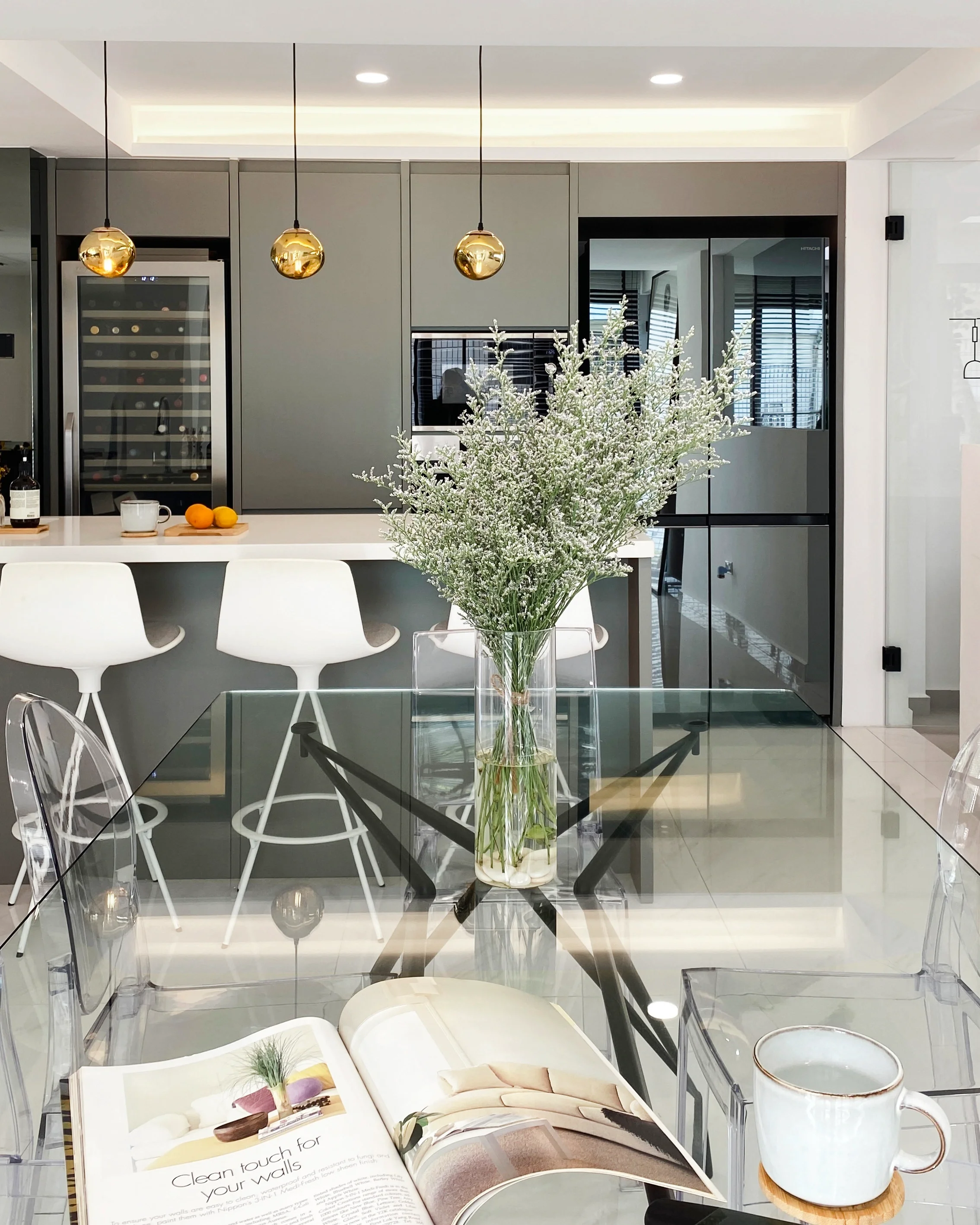 Modern kitchen with gray cabinets, white island with bar stools, hanging gold pendant lights, a glass dining table with a vase of white flowers, an open magazine, and a coffee mug.