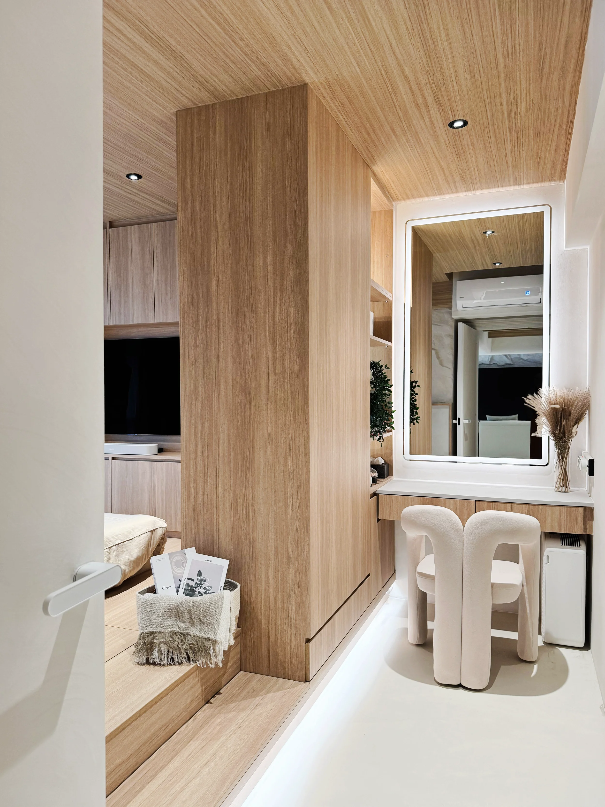 A modern hotel room with wooden paneling, a built-in desk with a mirror, a chair, a vase with dried flowers, a TV, and a bed partially visible.