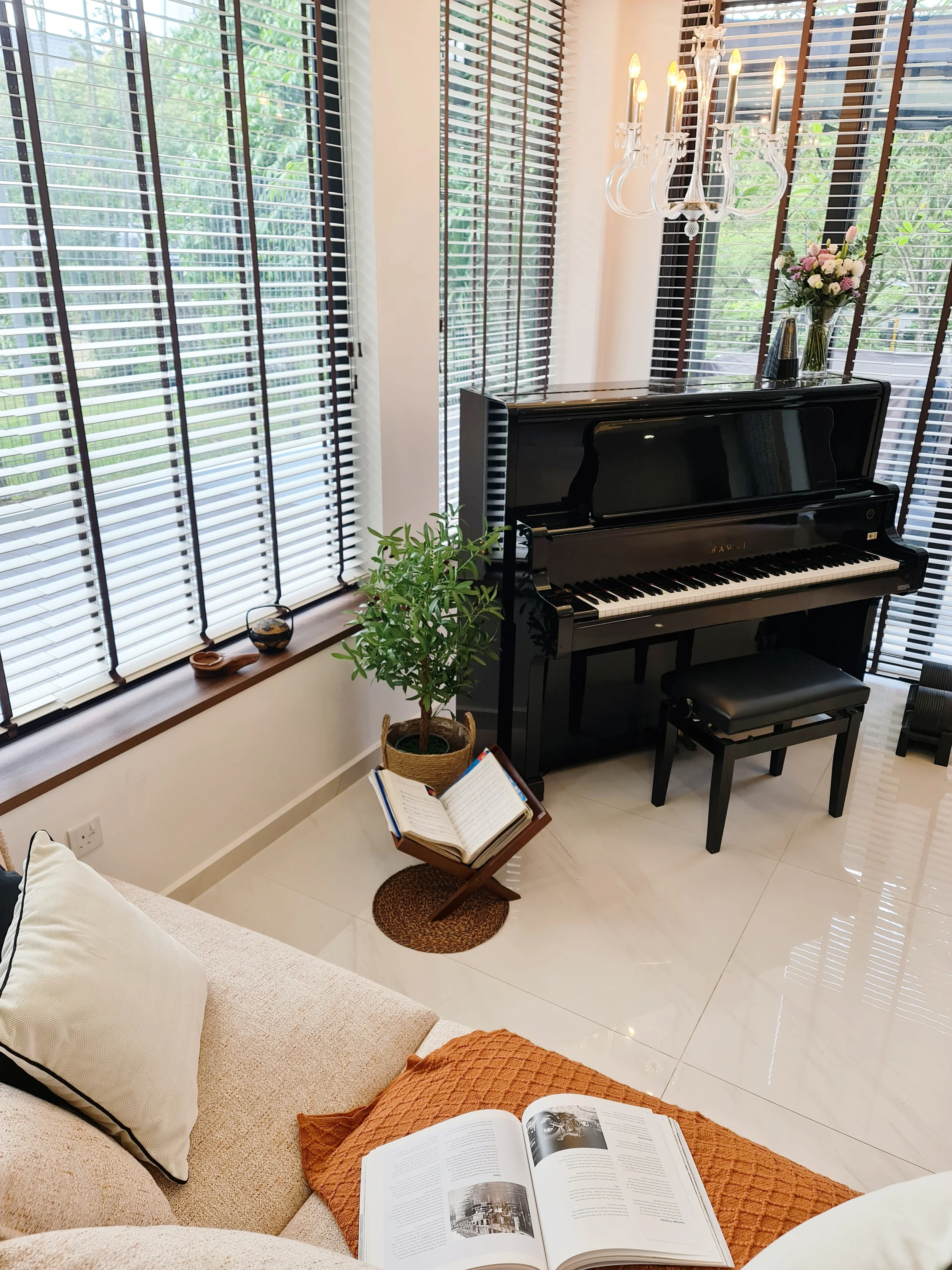 A cozy living room corner with a black upright piano near large windows with white blinds, a potted plant, an open book on a small wooden stand, a beige sofa with white and black pillows, a magazine on an orange blanket, a chandelier, and a vase of flowers.