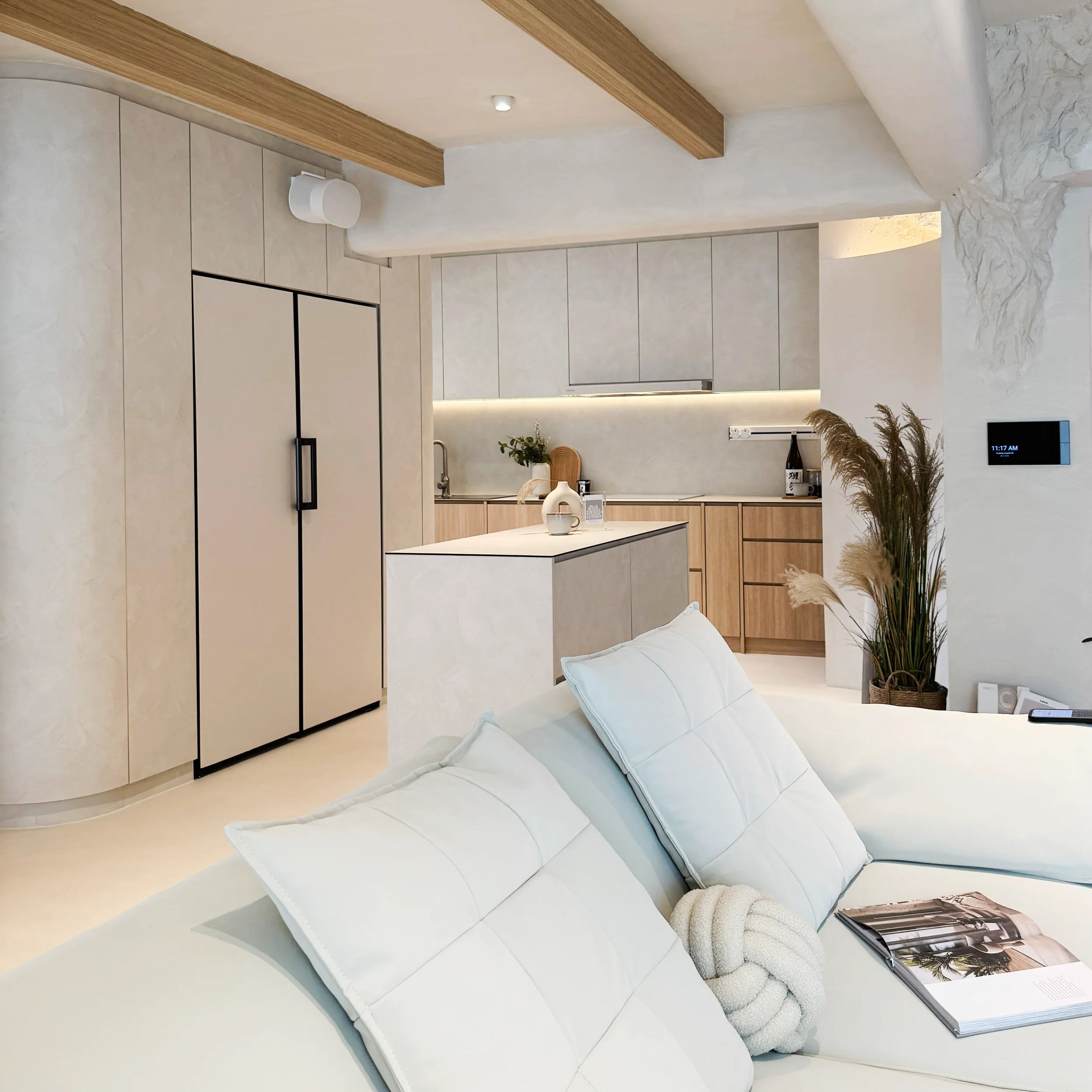 Modern living room with white sofa, pillows, and an open kitchen with light cabinets and wooden accents.