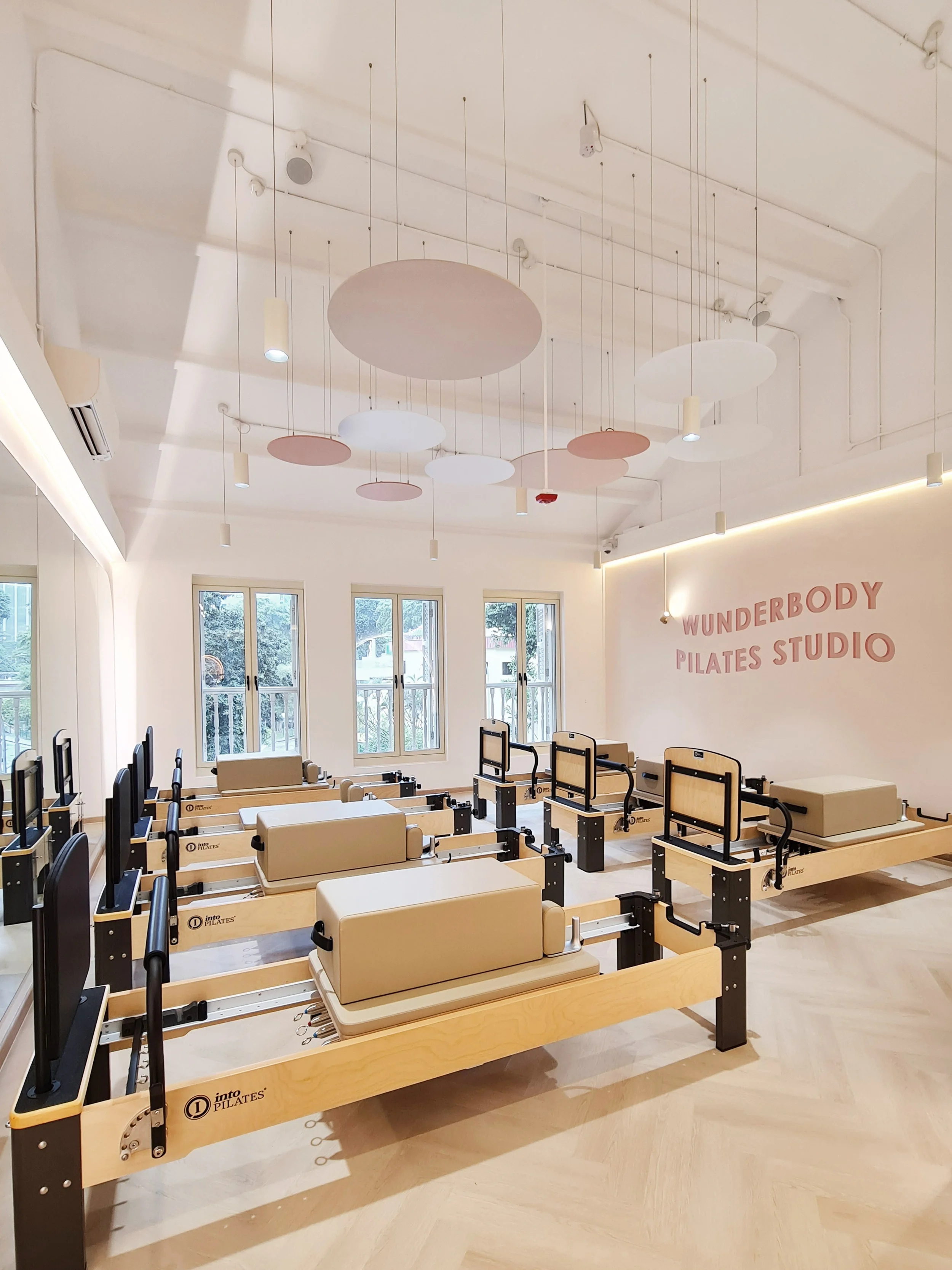 A Pilates studio with reformer machines arranged in rows, large windows, and a white wall with pink letters spelling 'WUNDERBODY PILATES STUDIO'.