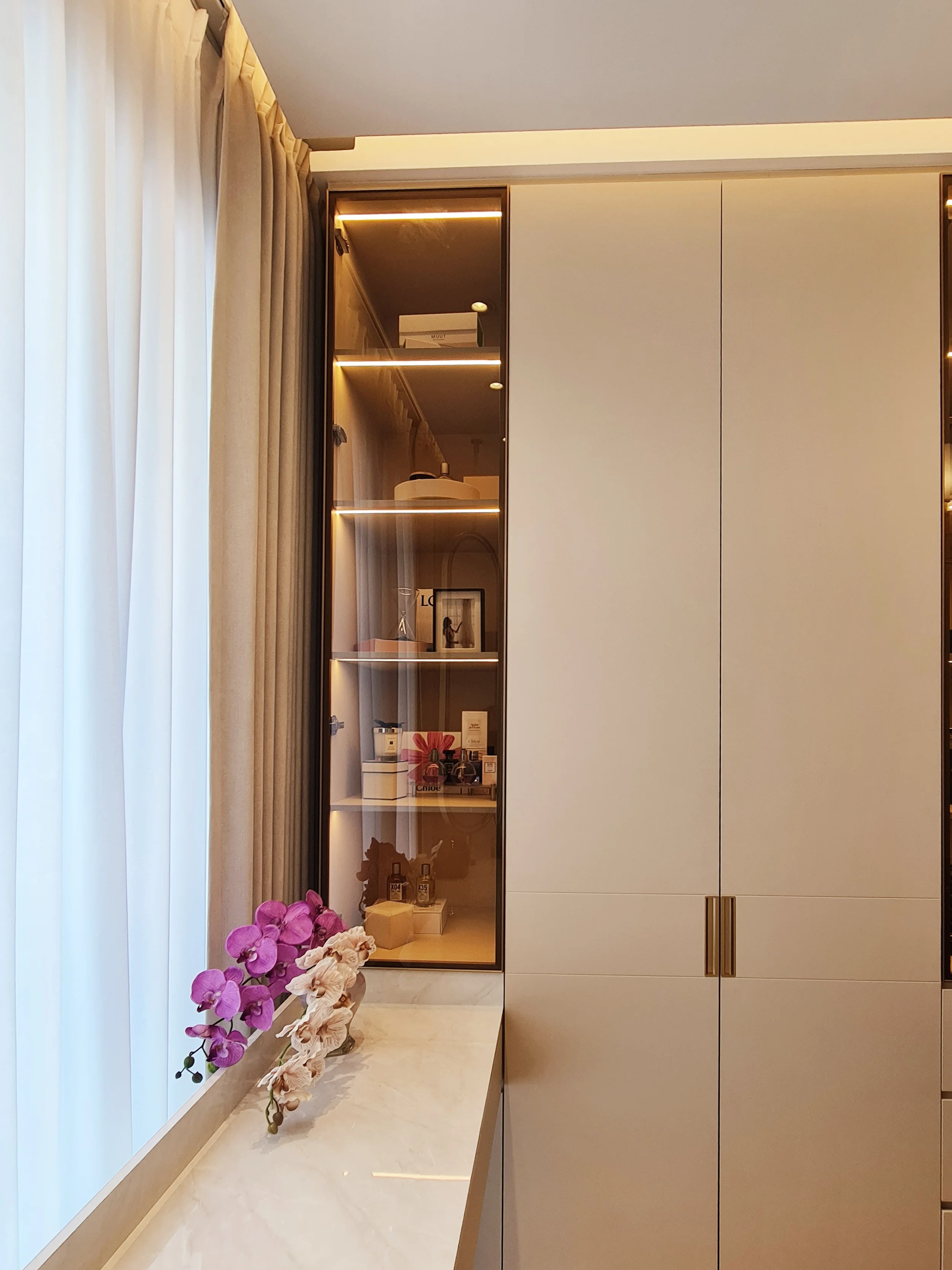 A white marble countertop with a pink and white orchid flower plant, next to a window with white curtains, and a tall glass display cabinet with shelves holding decorative items and cosmetics.