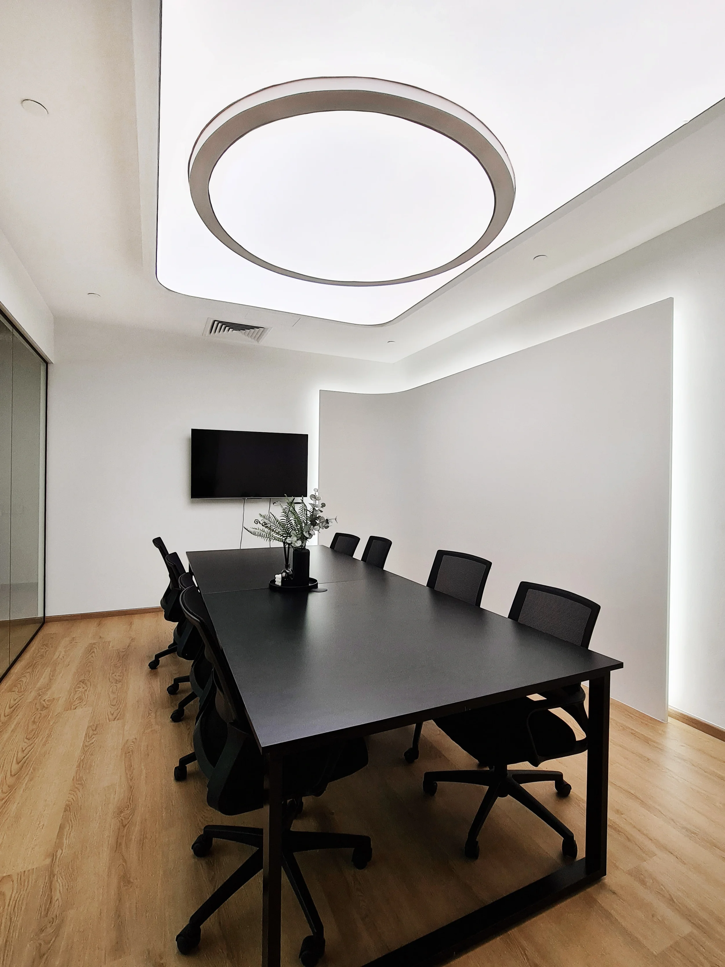 Empty modern conference room with a long black table, black office chairs, a wall-mounted TV, and a ceiling light fixture.